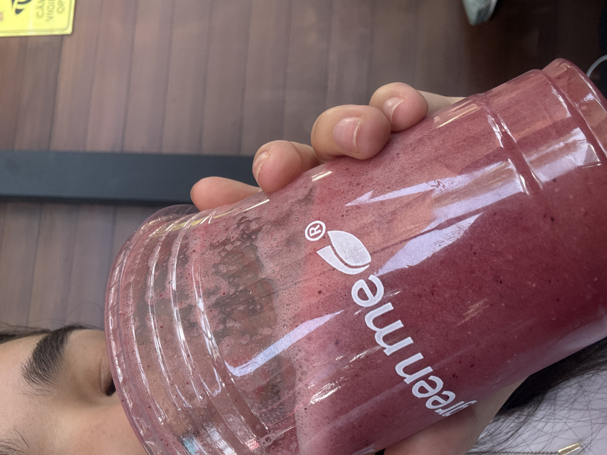 A person is holding a plastic cup with a red smoothie. The cup has the word "greenme" printed on it in white. The person's hand is visible, with their fingers wrapped around the cup. Part of the person's face, including their eye and eyebrow, is visible behind the cup. The background includes a wooden surface, a black bar, and a yellow sticker. There is insufficient information in the image to identify the location as Puebla, Mexico.