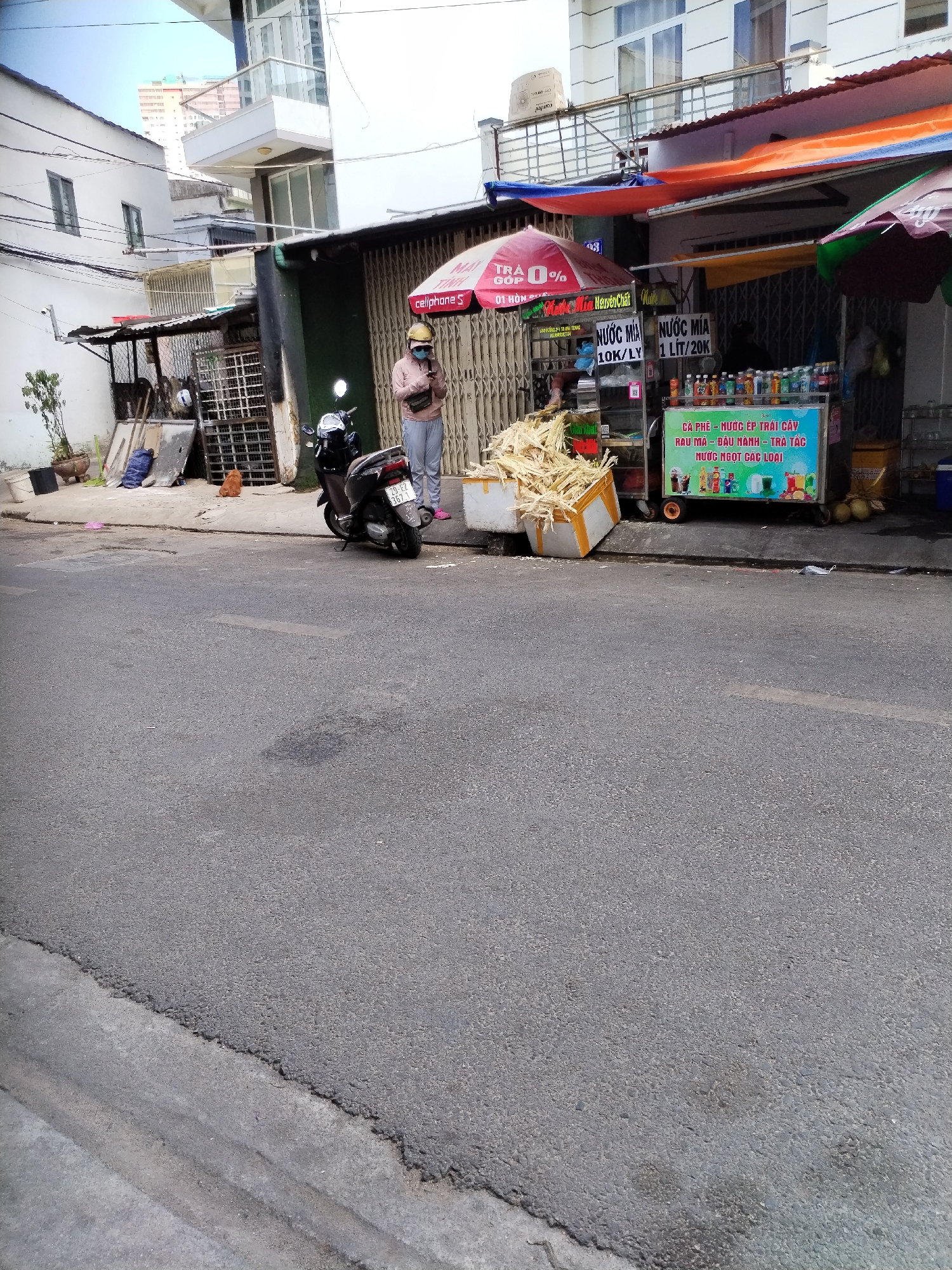 In Bac Nha Trang Ward, Vietnam, a street scene unfolds with a vendor standing beside a cart selling sugarcane juice and other beverages. A person, likely the vendor, wears a pink long-sleeved shirt, gray sweatpants, and a yellow helmet, with a mask covering their face. They are holding a mobile phone. A dark-colored motorcycle is parked to their left. The cart is adorned with colorful signs advertising various drinks, including "Nước mía" (sugarcane juice) at different price points. Stacked sugarcane stalks are visible in a white cooler box in front of the cart. The street itself is paved with asphalt, showing signs of wear. Buildings line the background, some appearing to be residential or commercial establishments, with awnings and metal grates visible. A red umbrella with "TRA Góp 0%" is open next to the vendor. A small, light brown dog is present on the sidewalk near some discarded items. The overall environment suggests a bustling street-level commerce.