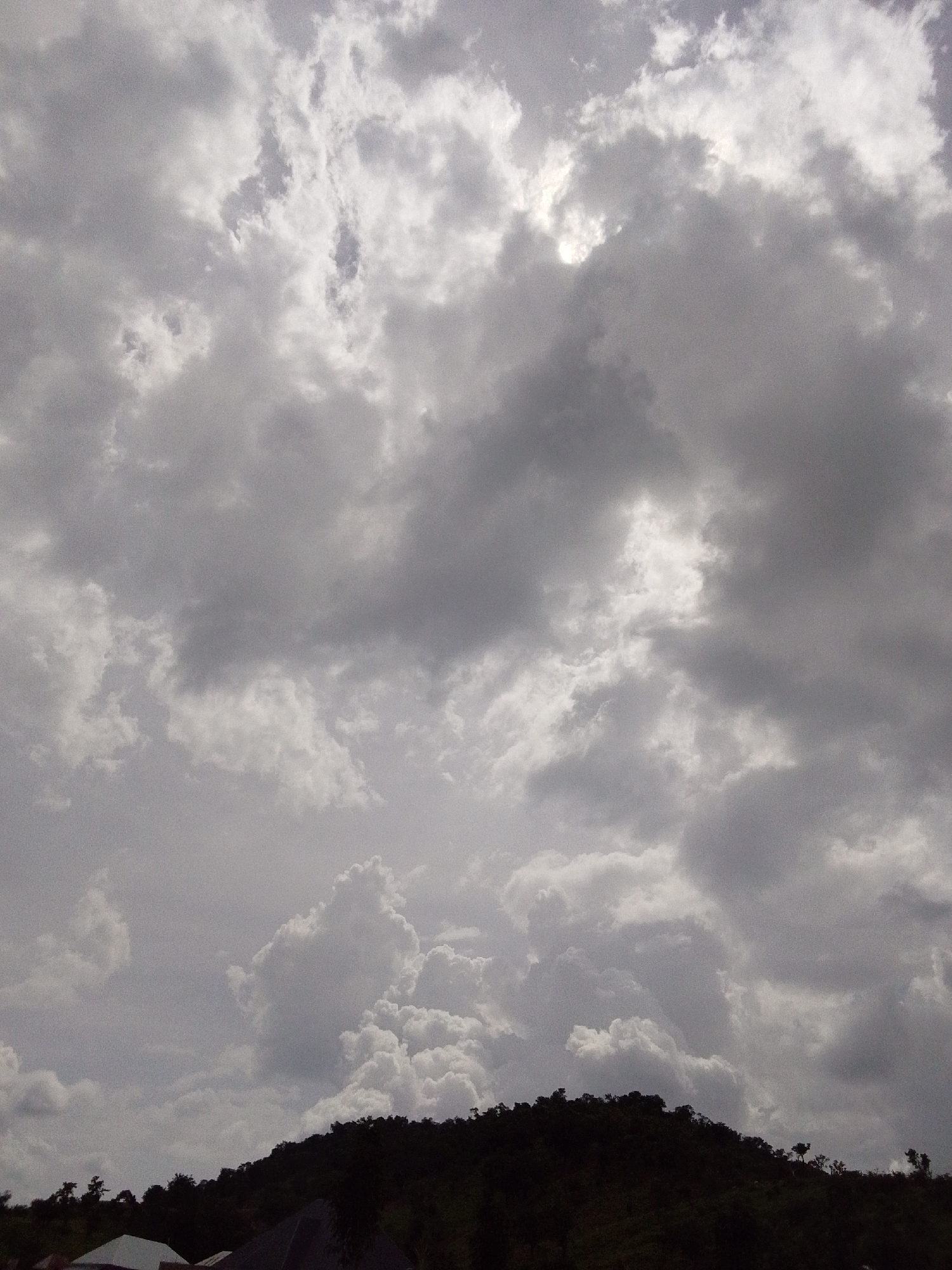 A low-angle, full shot captures a dramatic sky filled with heavy, grey, and white clouds. The clouds dominate the frame, with patches of brighter light suggesting the sun is attempting to break through. Below the tumultuous sky, a dark, tree-covered hillside forms the lower third of the image. Silhouetted against the sky, the foliage on the hill appears dense. In the bottom corners, the very tops of a few structures are visible, suggesting the presence of a settlement, possibly in the city of Serti, Nigeria. The overall mood is overcast and potentially foreboding, characteristic of an impending storm or heavy rainfall.