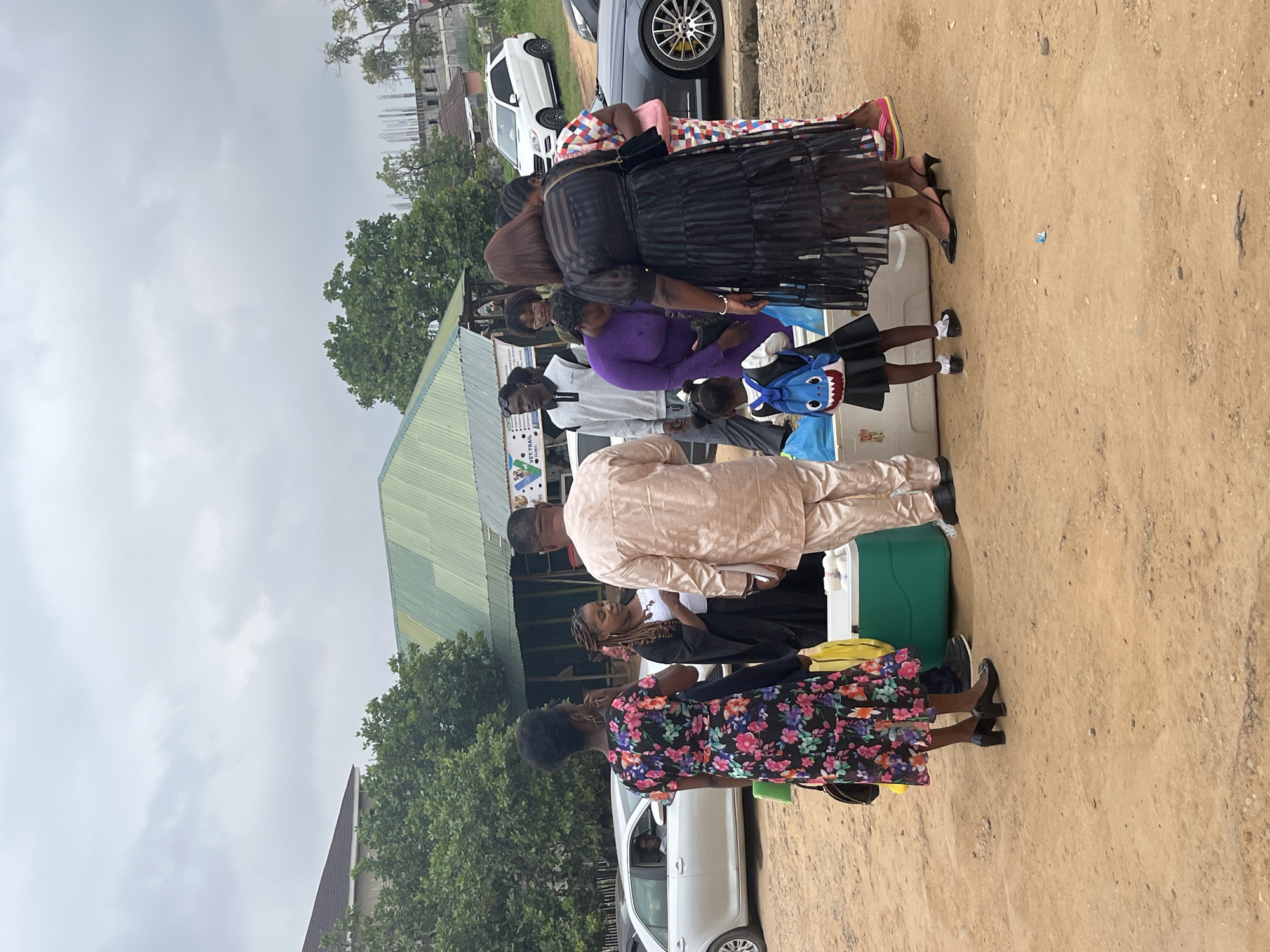 In Galadimawa Village, Nigeria, a group of people are gathered outdoors on a dirt lot near a building with a green corrugated metal roof. Several individuals, including women in dresses and a man in traditional attire, are interacting around a large green cooler and some white containers. A young girl wearing a backpack and a child in a dark dress are also present. In the background, cars are parked, including a silver sedan and a white SUV, with buildings and trees visible. The sky is overcast.