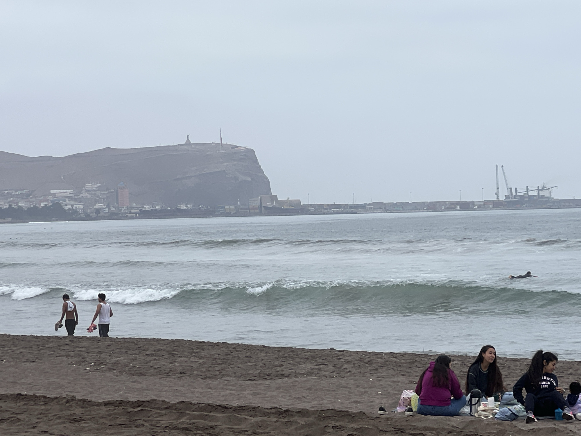 The image depicts a coastal scene in Arica, Chile, on a cloudy day. In the foreground, four people are gathered on a dark sand beach, seemingly having a picnic. Three individuals are seated, two women facing towards the viewer and one woman to the right facing away. Another person is seated to the far right, partially obscured. Objects such as bags, a thermos, and what appears to be food containers are scattered around them.
Further into the frame, two young men are walking towards the ocean from the beach. One carries a towel over his shoulder, and the other holds a pink object. The ocean displays moderate waves, and a lone surfer is visible in the distance.
The background reveals a cityscape built on the slopes of a large hill, with various buildings and structures. Atop the hill, a prominent statue of Christ the Redeemer is visible, a landmark of Arica. Beyond the city and the hill, industrial facilities with cranes and shipping containers are situated along the coastline, indicating a port area. The sky is overcast, casting a diffused light over the entire scene.