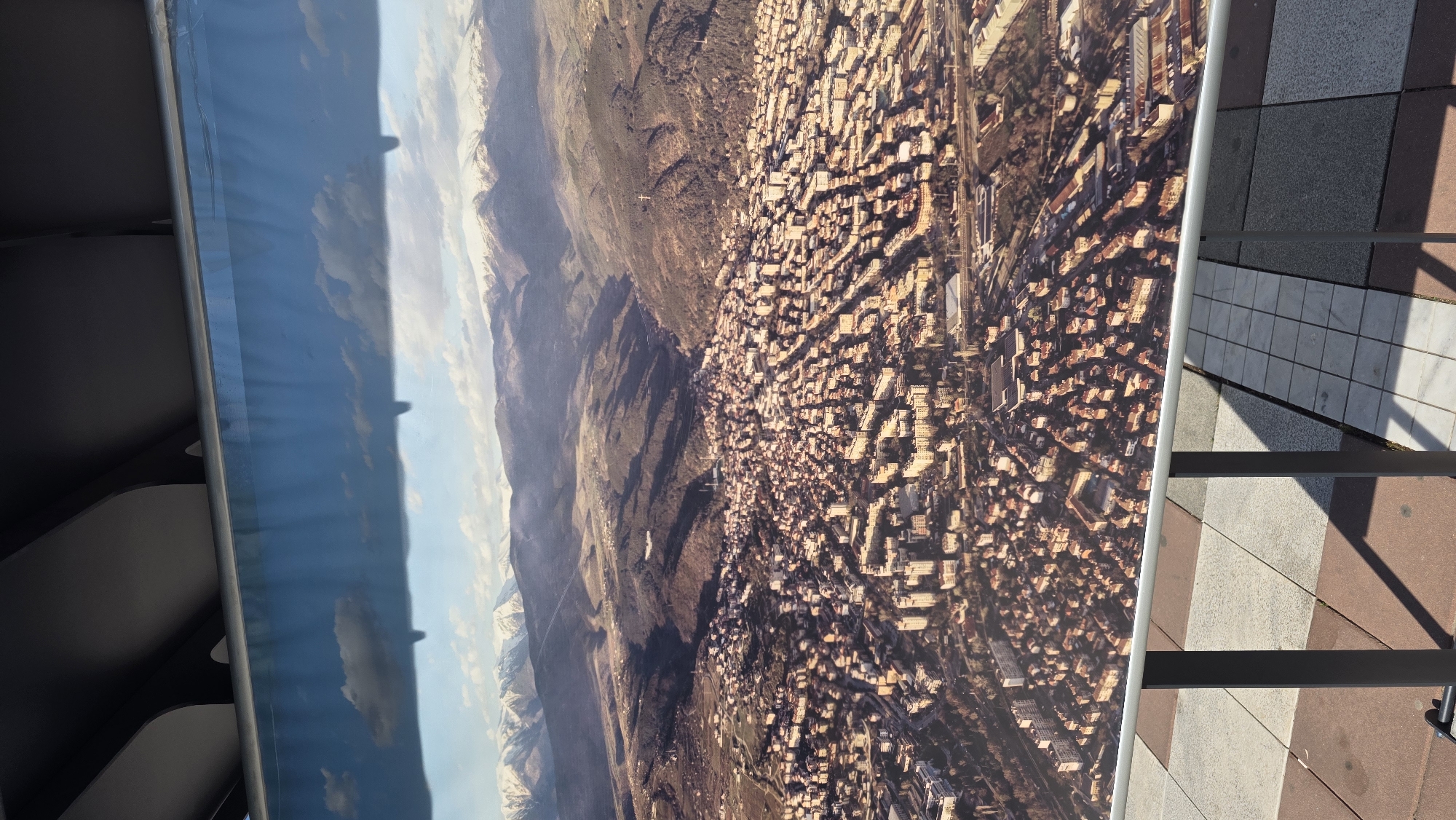 The image shows an aerial view of Blagoevgrad, Bulgaria, with the Rila mountains visible in the background. The city is densely populated with buildings, predominantly rectangular in shape and light brown in color. A network of roads and railway lines is visible throughout the urban area. To the left of the city, the mountainous terrain is mostly brown and barren. A blue sky with scattered clouds is present above the mountains. The image is displayed on a banner held by a metal frame, with a tiled sidewalk visible on the right.
