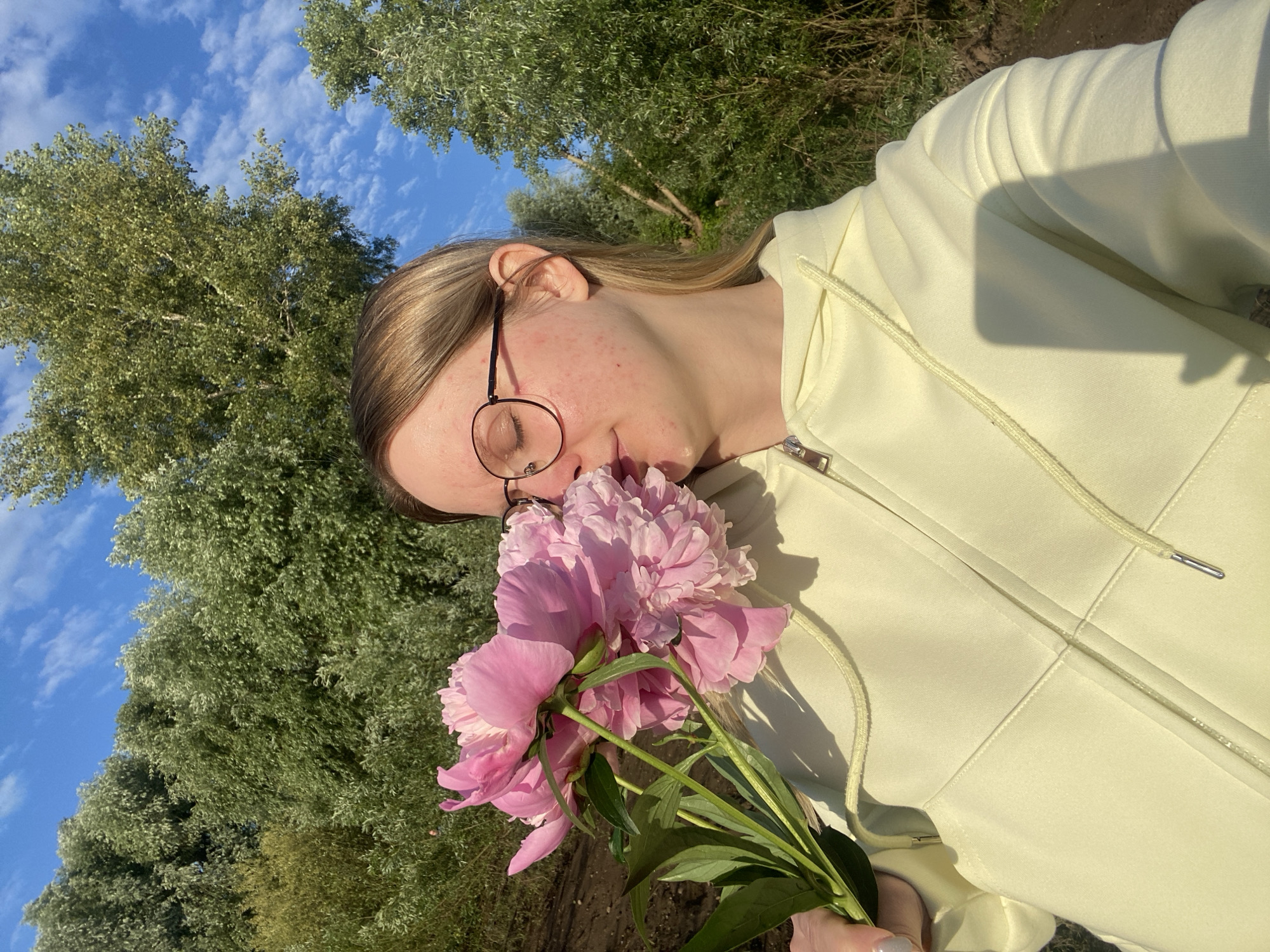 The image shows a young woman with blonde hair and glasses holding a bouquet of pink peonies to her face, seemingly smelling them. She is wearing a light yellow hooded jacket with a zipper. The background consists of lush green trees and a bright blue sky with scattered clouds, suggesting an outdoor setting. The ground appears to be a mix of dirt and grass. The lighting indicates it is daytime, possibly late afternoon due to the warm tones. The overall scene evokes a sense of nature and tranquility. The location is not explicitly identifiable as Chaikovsky, Russia, from the visual information alone.