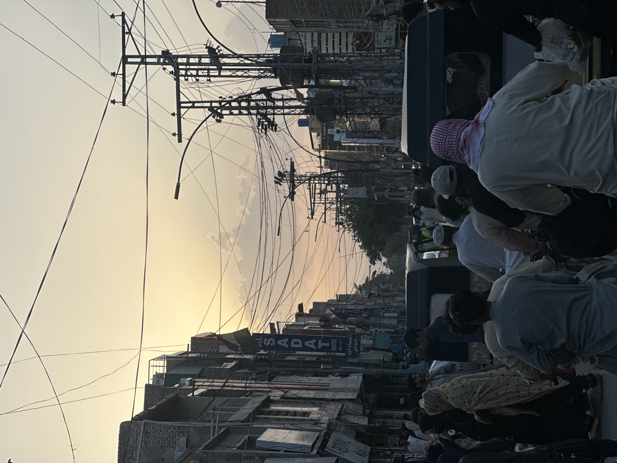 The image depicts an urban street scene in Quetta City Tehsil, Pakistan. Power lines crisscross above a densely packed street lined with buildings. A crowd of people is visible in the foreground, some appearing to be on a vehicle or platform. The sky is light, suggesting either dawn or dusk. Prominent signs for "SADAT HOTEL" are visible among the buildings. The scene seems busy and crowded.