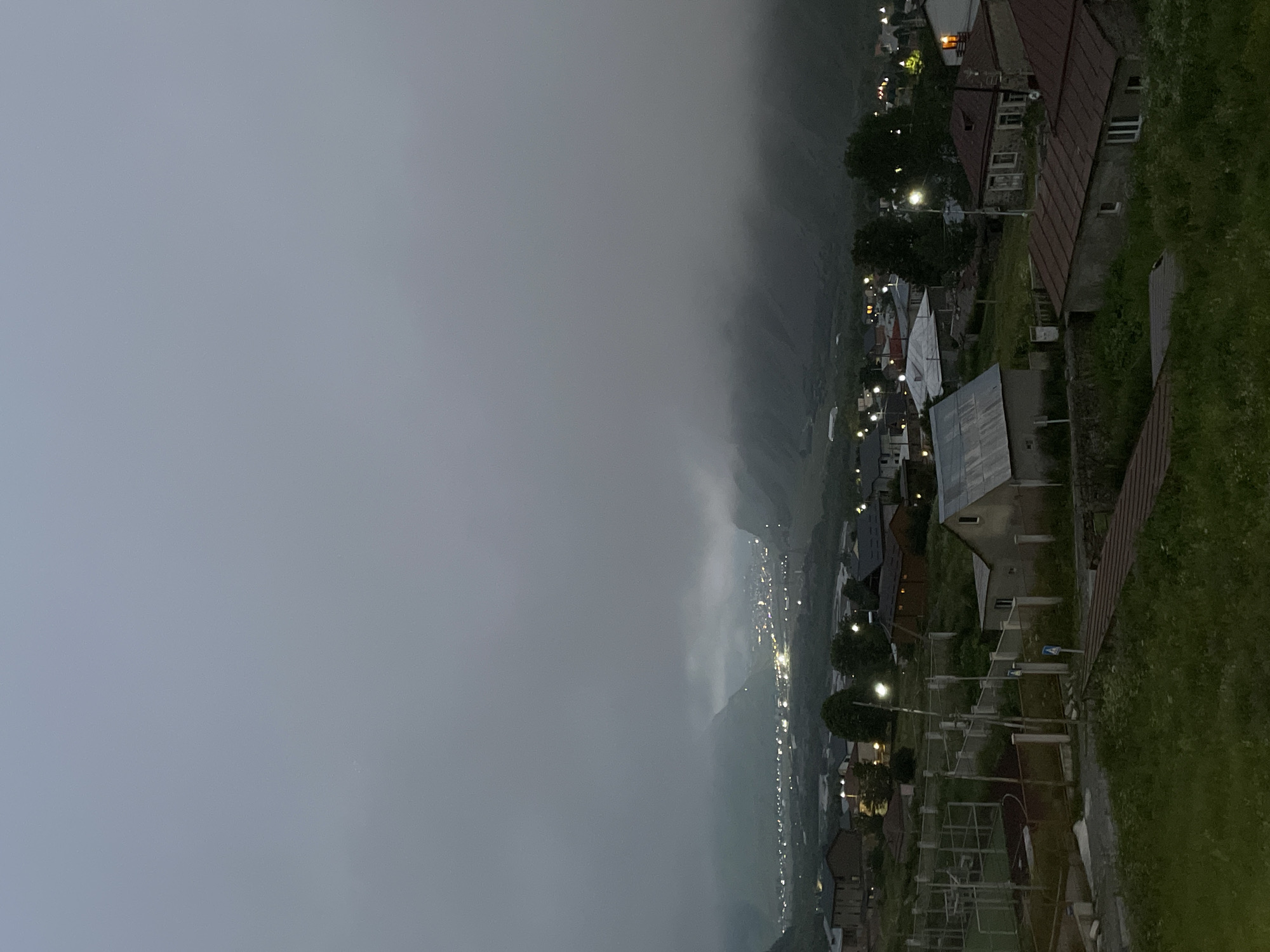 The image depicts a twilight scene in Stepantsminda, Georgia, characterized by low-lying clouds and mist obscuring the upper portions of the surrounding mountains. A small town is visible in the valley, with numerous lights illuminating the settlement, indicating it is either early morning or late evening. In the foreground, several houses with dark-colored roofs are situated on a grassy hillside. Some of these houses have visible windows, and one has a light on in an upper window. A sports field with fencing and a dark red surface is also present on the left side of the foreground. The overall atmosphere is somewhat hazy and subdued due to the ambient light conditions and the mist.