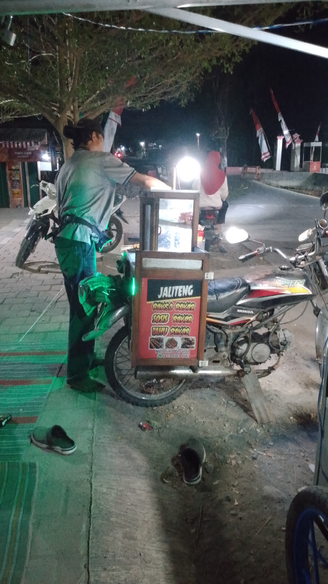 At night in Karanglangu, Indonesia, a person wearing a grey shirt and dark pants stands behind a mobile food cart attached to a motorcycle. The cart displays a sign for "JALITENG" and lists food items such as "BAKSO BAKAR," "SOSIS BAKAR," and "TAHU BAKAR," along with a phone number for orders. The motorcycle is a dark color with "HONDA" branding visible. In the background, other motorcycles and a person wearing a red headscarf are visible. Flags, possibly national flags of Indonesia, are mounted on poles in the distance. The scene appears to be on a paved area, possibly a street or sidewalk, with some vegetation and a tree present. Several pairs of footwear are scattered on the ground.