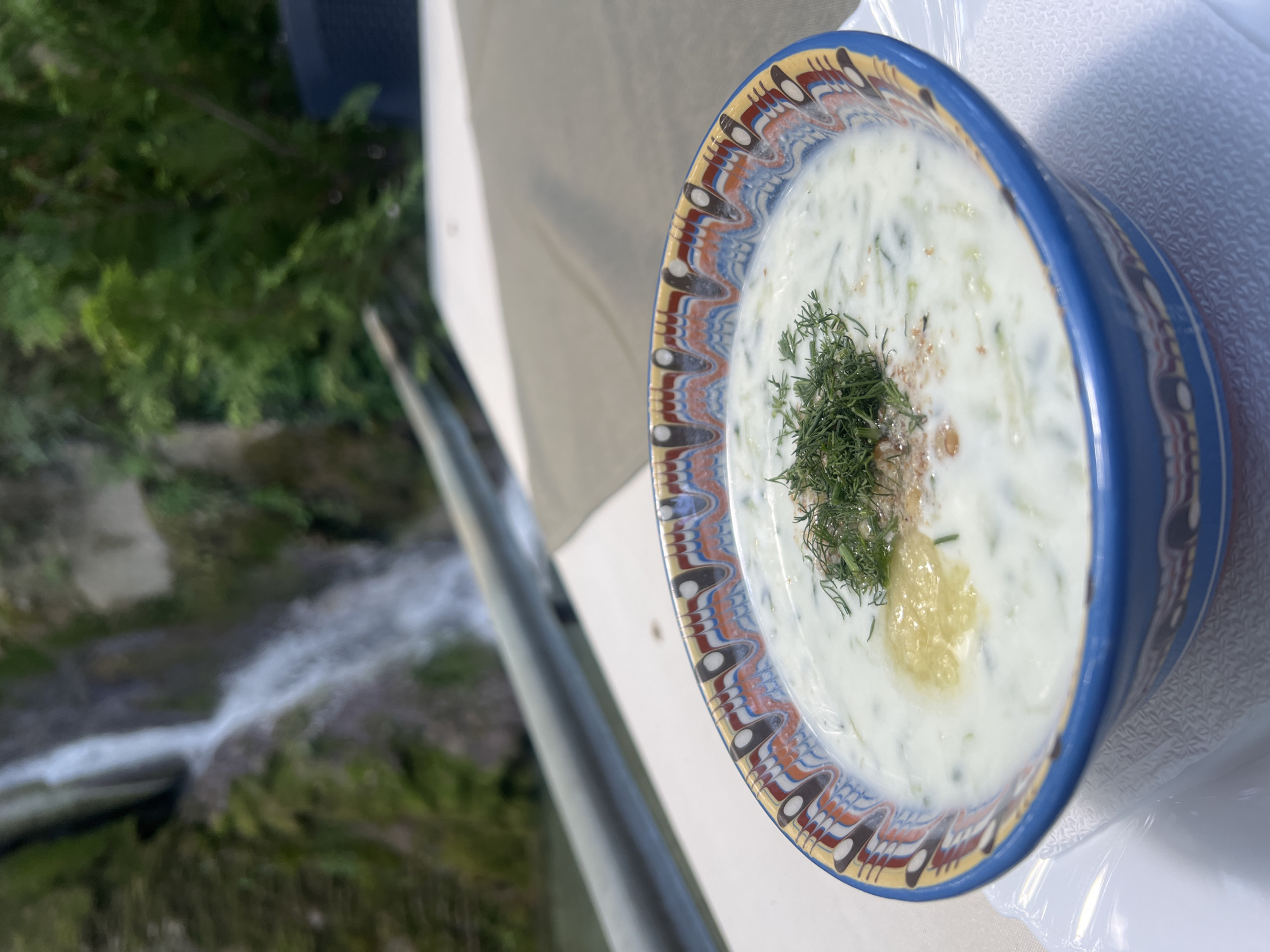 A close-up, slightly angled shot captures a bowl of cold soup, likely a refreshing yogurt-based dish with cucumber and dill, garnished with more dill and a dollop of yellow dressing. The soup is served in a vibrant, ethnically patterned blue bowl, resting on a white textured napkin and a white plate. The background is softly blurred, revealing a natural landscape with a cascading waterfall, lush green foliage, and a metal railing. The overall setting suggests an outdoor dining experience, possibly at a restaurant or cafe near a natural landmark in Bachkovo, Bulgaria, offering a picturesque backdrop to the meal.