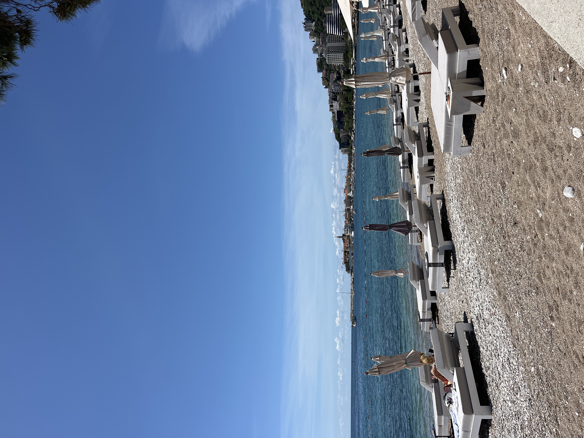 The image shows a beach scene in Budva, Montenegro. The view is along the coastline, showing a line of white beach loungers and umbrellas on a pebble beach. The Adriatic Sea is adjacent to the beach, and in the distance, buildings of Budva are visible along the coast. The sky is a clear blue, with a few wispy clouds. A person is lounging on one of the beach chairs.