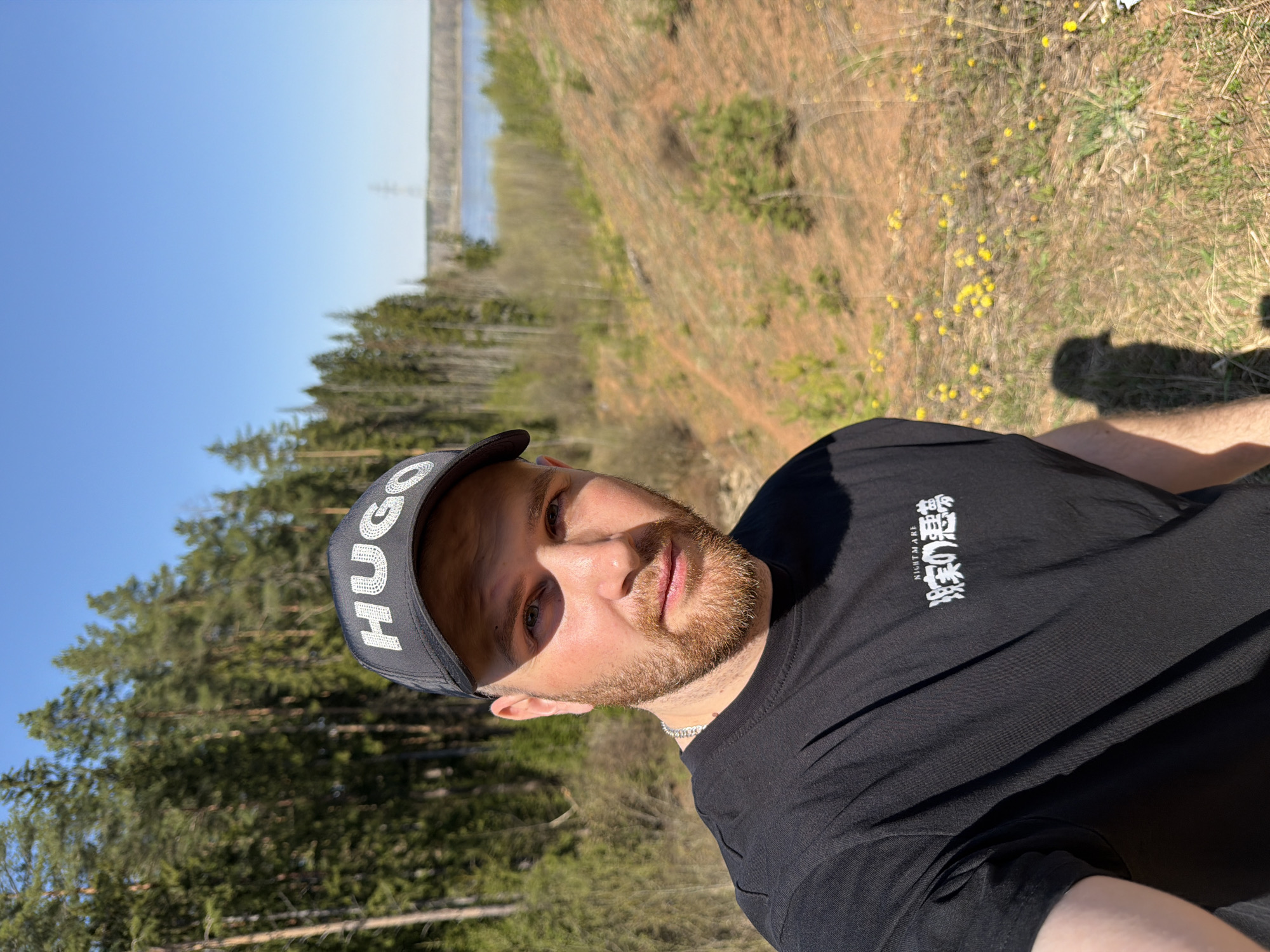 A selfie shot of a man in an outdoor location. He is wearing a black t-shirt with Japanese writing on it and a black baseball cap with the brand "HUGO" on it. He has short, light brown hair and a beard. The background includes trees, a grassy area with small yellow flowers, and a body of water in the distance. The location is Priokamsky, Russia. The image is taken in daylight, with clear sky.