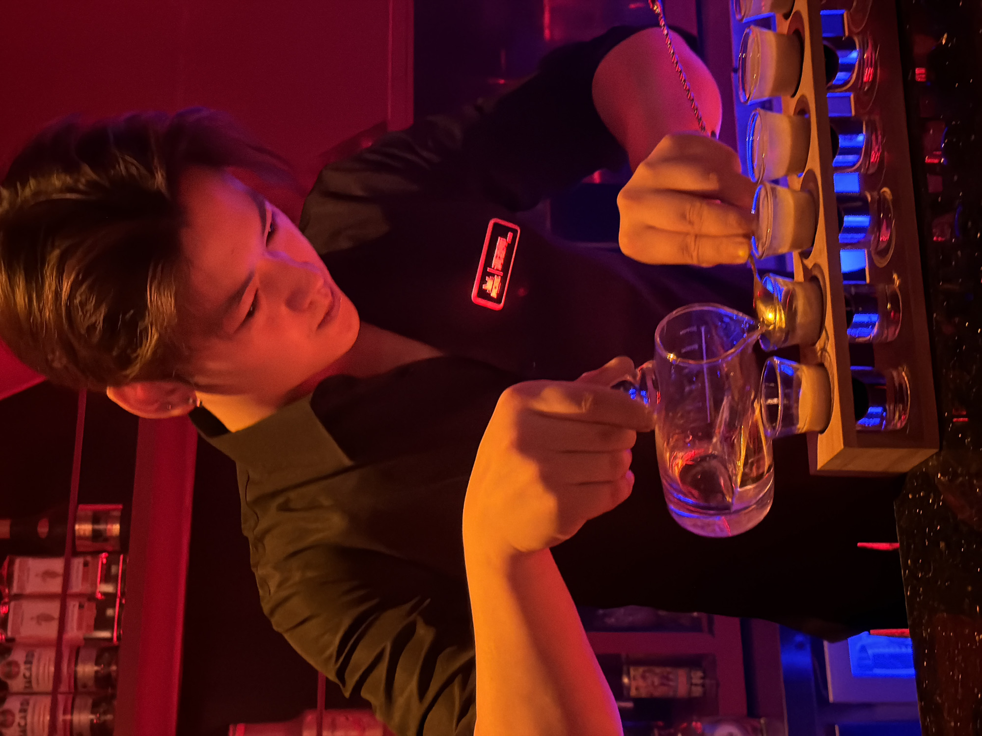 A young male bartender, wearing a black uniform shirt with a red illuminated logo on the left chest, is preparing shots behind a dimly lit bar. He is in the process of pouring a liquid from a decanter into a row of small shot glasses. His head is bent downwards, focusing intently on his task. The background features shelves stocked with bottles, illuminated by red and blue lights, suggesting a nightlife establishment. The overall atmosphere is dark and intimate. The location is identified as Jiangbeicheng Subdistrict, China.