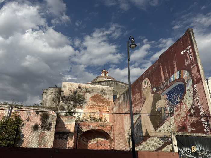 The image captures an urban street scene in Puebla City, Mexico, featuring a prominent mural on a weathered wall, old architecture, and a partially cloudy sky.
In the midground, a large, reddish-brown and pinkish-orange wall dominates the right side of the frame, angled sharply towards the viewer. This wall is adorned with a vibrant, though faded and peeling, street art mural. The mural depicts a stylized, light-skinned human-like figure with a complex, brain-like internal structure visible within its head. The figure appears to be interacting with a large, abstract, colorful object that resembles a fragmented, perhaps mechanical, device or a surreal landscape. The colors in the mural include blues, greens, yellows, and oranges, contrasting with the dark red of the wall. Above the mural, at the top right corner of the wall, the text "PENSION 24 HRS" is visible in white and black lettering. Graffiti is also present at the base of this wall, though largely illegible.
To the left of the mural, and partially obscured, stands a black lamppost with a visible light fixture. Behind the lamppost and the mural wall, a much older, decaying stone structure rises, showing signs of significant age with crumbling plaster, exposed brick, and abundant wild vegetation growing on its surfaces. At the very top of this background structure, a red-domed roof, typical of a church or colonial building, is visible against the sky. A chain-link fence runs along the bottom of the older walls.
The sky above is a mix of deep blue patches and scattered, fluffy white and grey clouds, suggesting a partly cloudy day. The overall lighting indicates daytime. The scene evokes a sense of historical depth combined with contemporary urban art, reflecting the character of an old city.