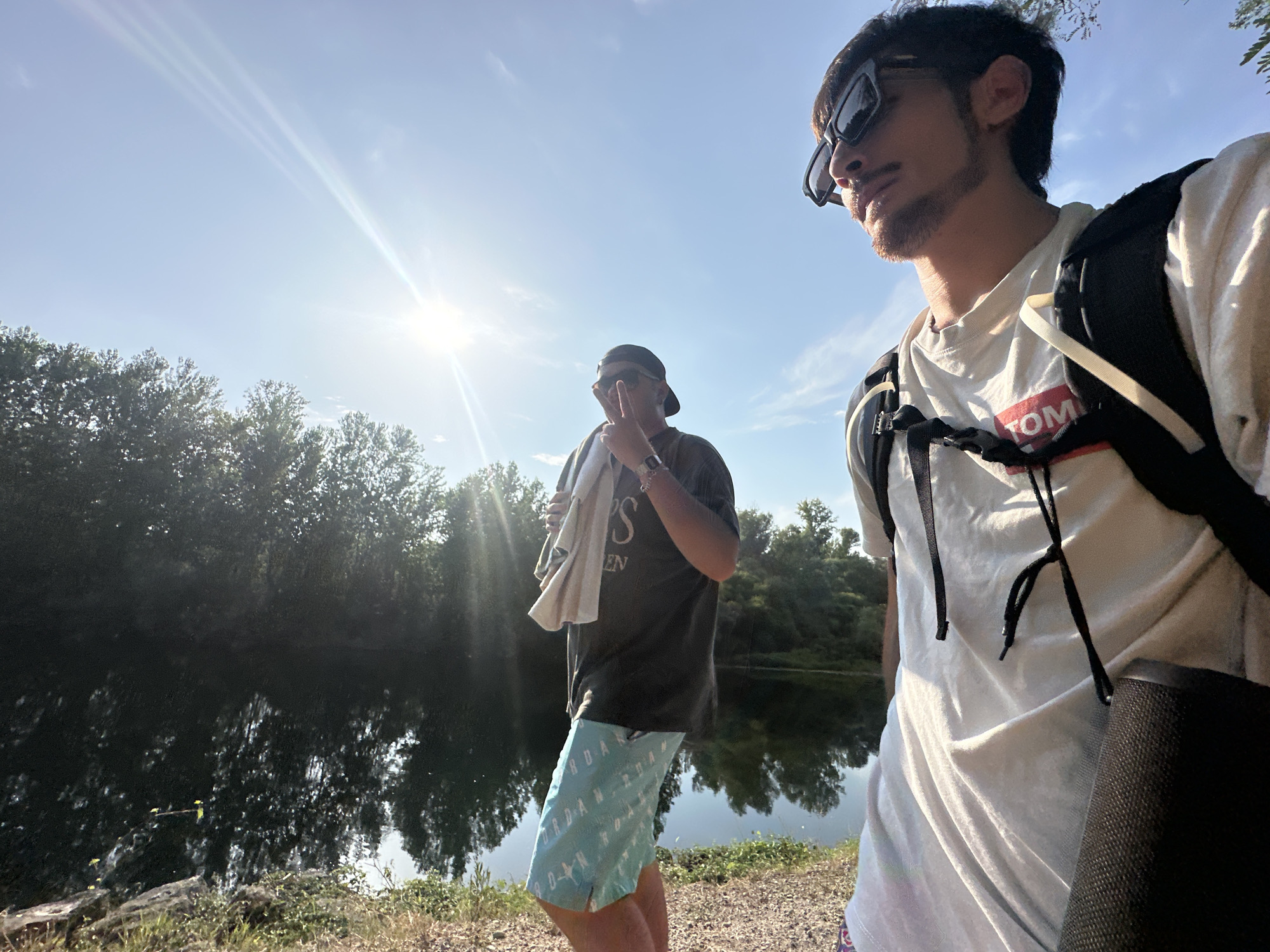 The image shows two young men standing by a body of water, likely the Oglio River in Rudiano, Italy, on a sunny day. The man in the foreground, wearing sunglasses and a white t-shirt with a black backpack, is looking to the right. The second man, partially obscured by the first, is wearing a dark t-shirt, a baseball cap, sunglasses, and light blue patterned shorts. He is holding a towel over his shoulder and making a peace sign with his fingers. The background features trees and their reflections in the water under a bright, sunny sky with lens flare. The ground is covered with gravel and vegetation.