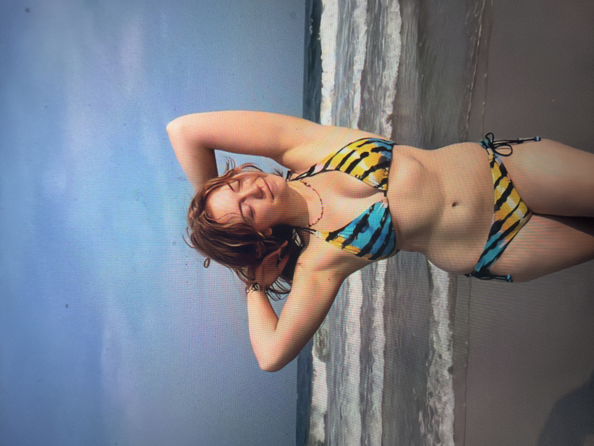 A young woman with reddish-brown hair and fair skin stands on a sandy beach, facing forward with her eyes closed and a smiling expression. Her arms are raised, with her hands behind her head. She is wearing a two-piece bikini featuring a pattern of blue, yellow, and black stripes or swirls. A thin, dark red beaded necklace is visible around her neck, and a dark-colored bracelet is on her right wrist. In the background, white-capped waves of the ocean break on the shore, transitioning into a light blue sky with some visible cloud formations. The entire image exhibits a prominent pixel grid pattern and scattered small, dark specks, indicative of a photograph taken of a digital screen. There are no identifiable features within the image to confirm its specific geographical location as Shumen, Bulgaria; the depicted scene is a coastal environment.