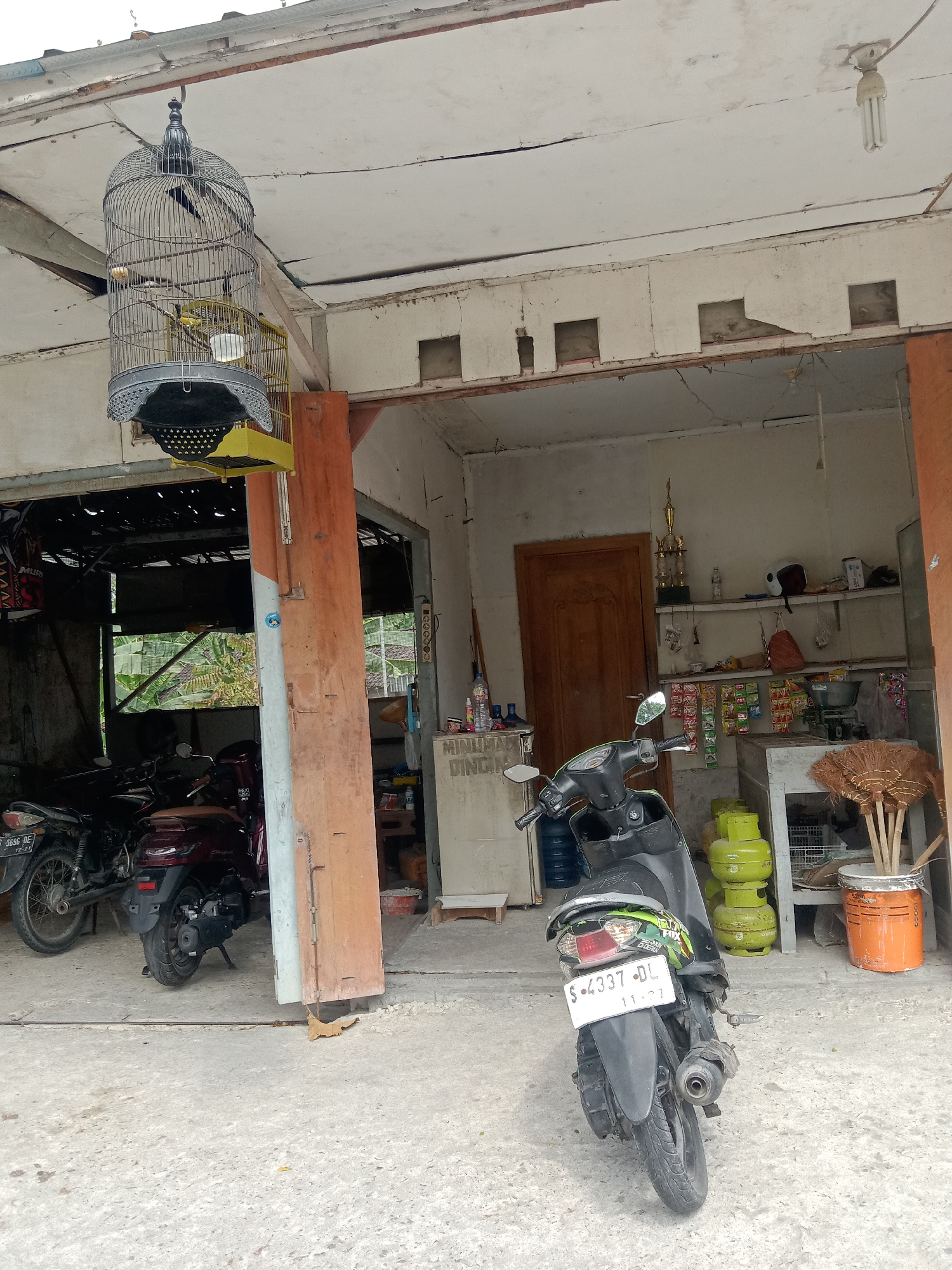 This is a medium shot, eye-level, outdoor photograph taken in a residential area in Siwalan, Indonesia. The scene depicts a covered porch area with several motorcycles parked in front of an open doorway leading into a building. A birdcage containing a small bird is hanging from the ceiling of the porch. To the right of the doorway, shelves are visible, stocked with various items including snacks, cleaning supplies, and gas cylinders. A broom stands upright in an orange bucket. The architecture suggests a modest, possibly rural, dwelling. The overall impression is one of everyday life in a small Indonesian town.