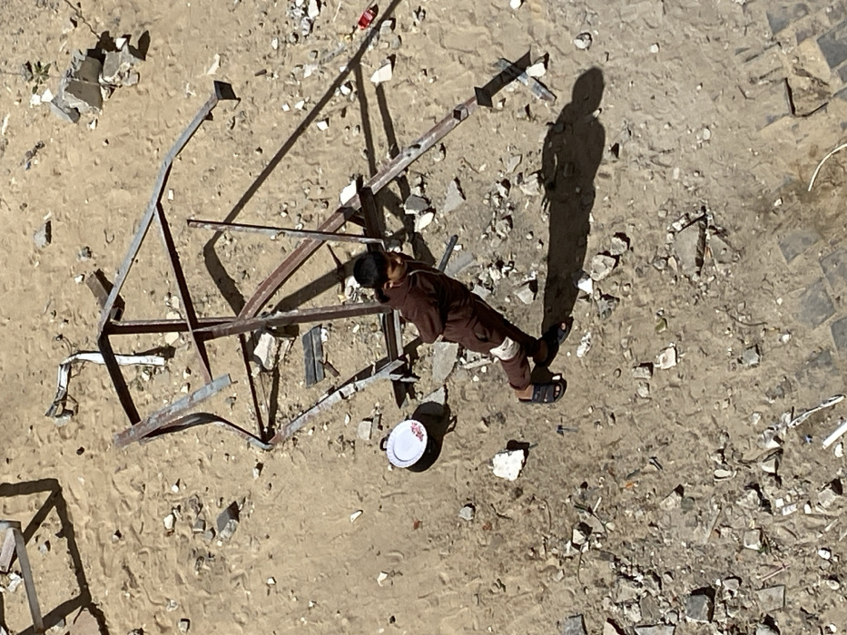 An overhead view in Khan Yunis, Palestinian Territory, depicts a young boy standing amidst sandy, debris-strewn ground. The boy, wearing a brown tracksuit and dark sandals, stands with his back to the viewer, his head turned slightly to his right, looking upwards. His arms are bent, with hands seemingly clasped behind his back. To his left, a white enamel plate with a floral pattern rests on the ground. Immediately behind him and extending across the upper portion of the frame are several pieces of twisted, rusted metal rebar and structural components, casting long shadows. The ground is a mix of light brown sand and dirt, heavily littered with irregular fragments of concrete and other building materials. Sunlight illuminates the scene from the upper left, casting distinct shadows.