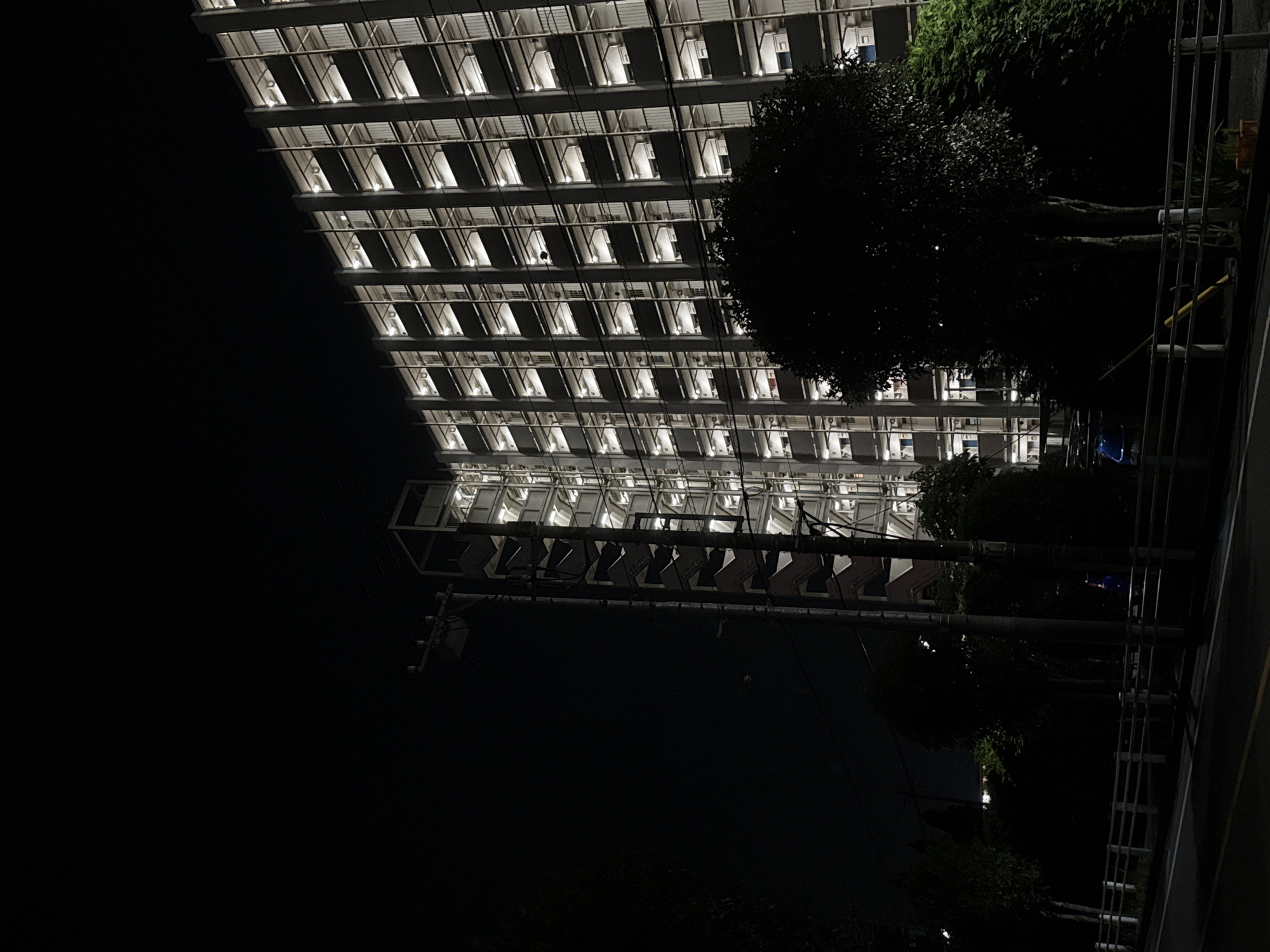 In Yachiyo, Japan, at night, a tall, multi-story apartment building is brightly lit with many windows illuminated. The building has a facade of repeating balconies or open areas. To the left of the building is an outdoor staircase structure, also visible with lights. In the foreground, the scene is dark with silhouetted trees and shrubbery obscuring the lower parts of the building. A road and a white railing are visible in the lower portion of the image. A utility pole with wires is situated between the street and the apartment building.