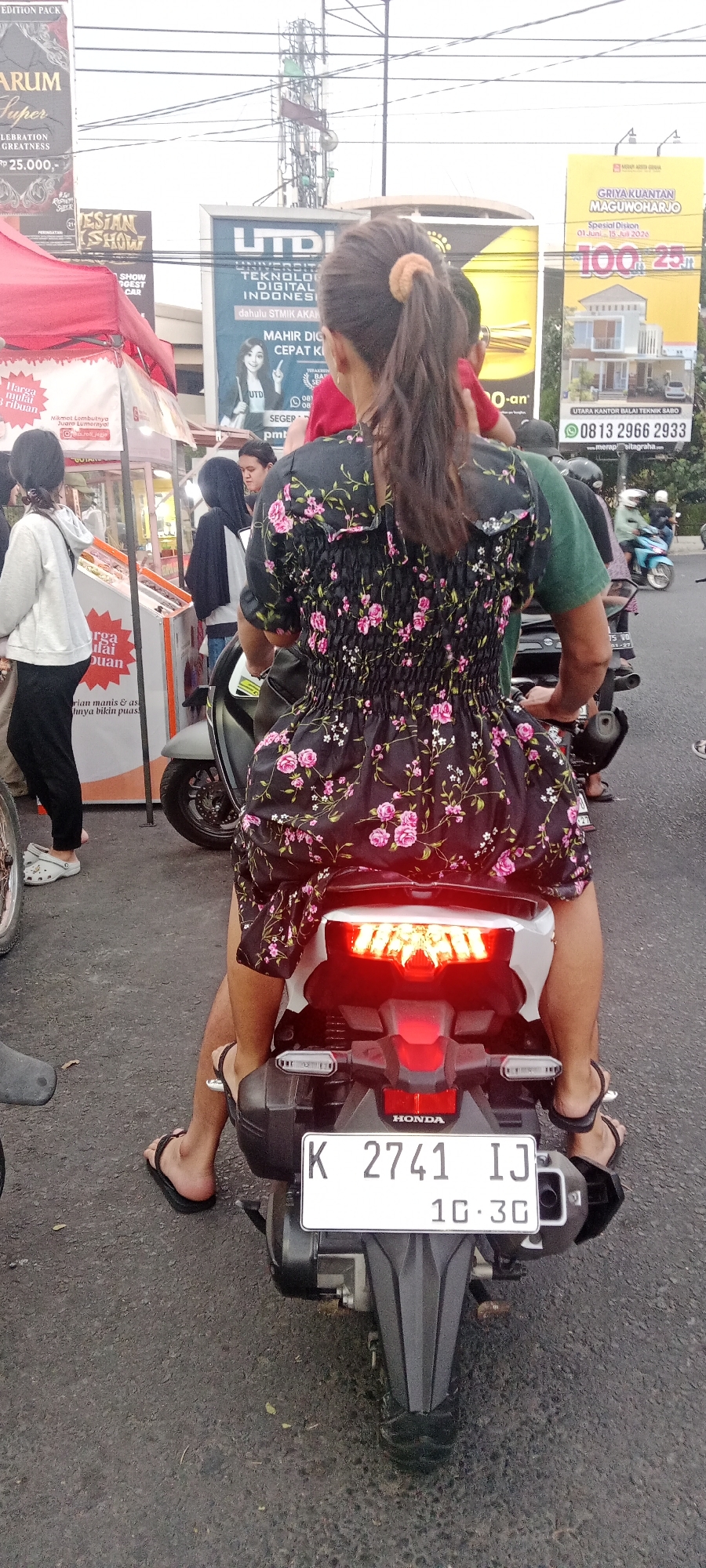 In the city of Ngemplak, Indonesia, on a paved street, a person wearing a black floral dress is sitting on the back of a motorcycle. They have dark hair pulled back in a ponytail with a light-colored scrunchie. The motorcycle has a white license plate that reads "K 2741 IJ" and "10.3G". The motorcycle's brake lights are illuminated. In the background, there are several billboards, a food stall under a red tent, and other people on motorcycles. One billboard is for "UTDI Universitas Teknologi Digital Indonesia," another promotes "Griya Kuantan Magunoharjo," and a third displays "ESTAN SHOW." The environment suggests a busy street scene with commercial activity.