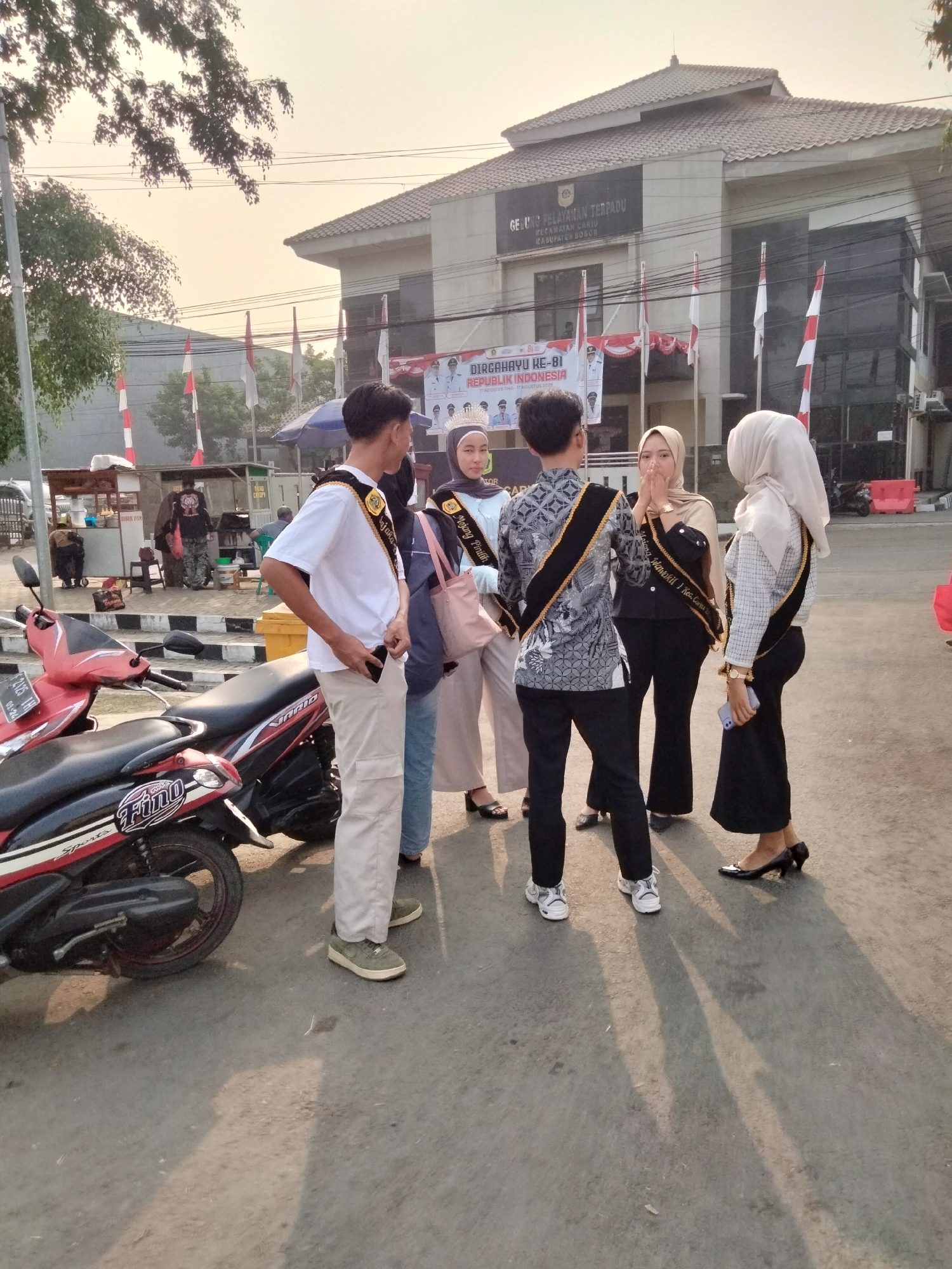 In Cariu, Indonesia, outside a building with a tiled roof and the signage "GEDUNG PELAYANAN TERPADU KECAMATAN CARIU KABUPATEN BOGOR", several young adults are gathered. The building appears to be a government office. Two motorcycles are parked in the foreground. The group consists of four individuals, two males and two females, all wearing sashes with gold trim and black lettering. The males are dressed in formal attire with sashes, while the females are in hijabs and elegant clothing, also with sashes. One female is holding a pink bag, and another is holding a smartphone. A banner celebrating "DIRGAHAYU KE-81 REPUBLIK INDONESIA" is visible on the building's facade. The scene is illuminated by the warm, low light of what appears to be either early morning or late afternoon.
