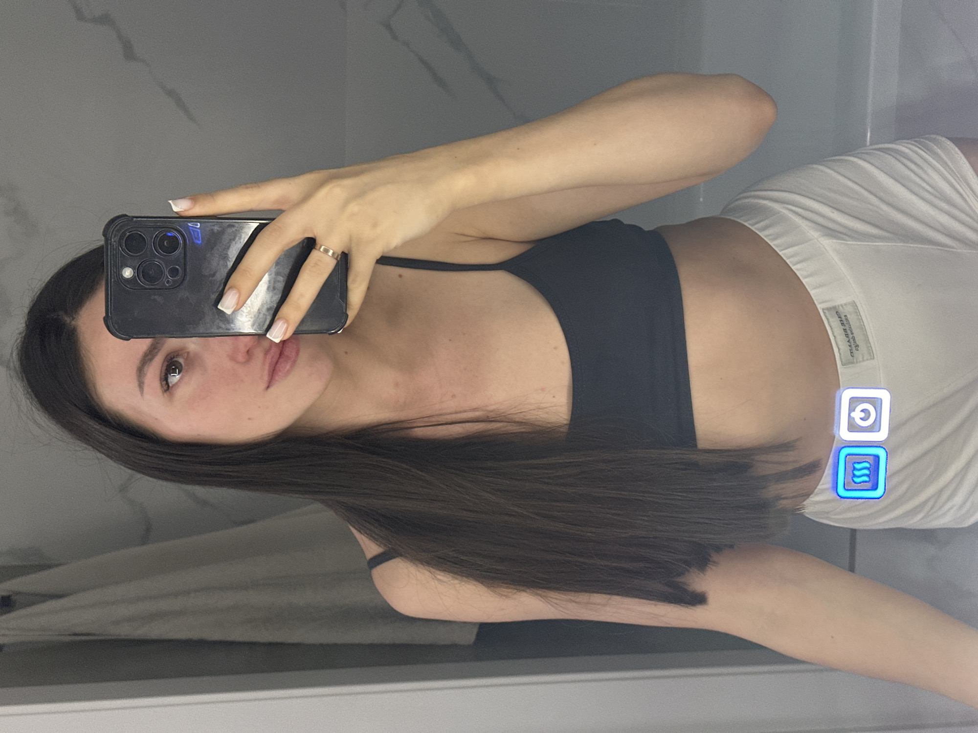 A woman with long dark hair, wearing a black sports bra and white shorts, takes a selfie in a bathroom. She is holding a black smartphone in her right hand, with her left hand resting on her hip. The background shows a tiled wall and a towel hanging on the left. Two illuminated icons, one depicting wavy lines and the other a power symbol, are visible on her shorts. The location is not specified as Эстачи, Russia.