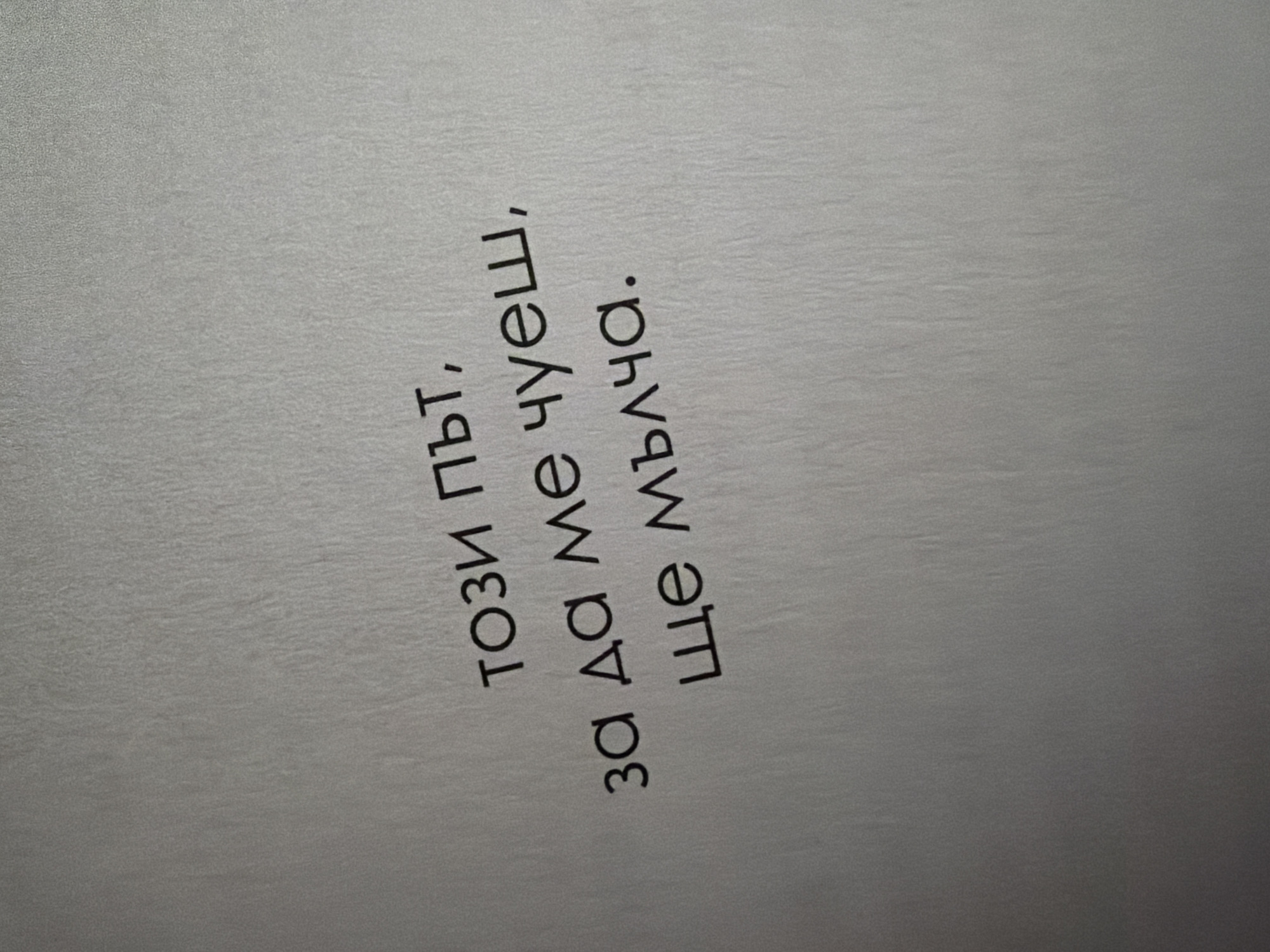 The image displays a close-up of text written on a light-colored, textured surface, likely paper. The text is in Bulgarian and is oriented diagonally from the bottom left to the top right of the frame. The words are in black, sans-serif font. The text reads: "този път, за да ме чуеш, ще мълча." which translates to "this time, so you can hear me, I will be silent." The background is a plain, uniform gray with subtle vertical textural lines, suggesting it is a document or a similar printed material. There are no people, animals, or any other objects visible in the image. The lighting appears to be ambient, creating no harsh shadows. There is no specific geographical location in Pazardzhik, Bulgaria, that can be identified from this image.