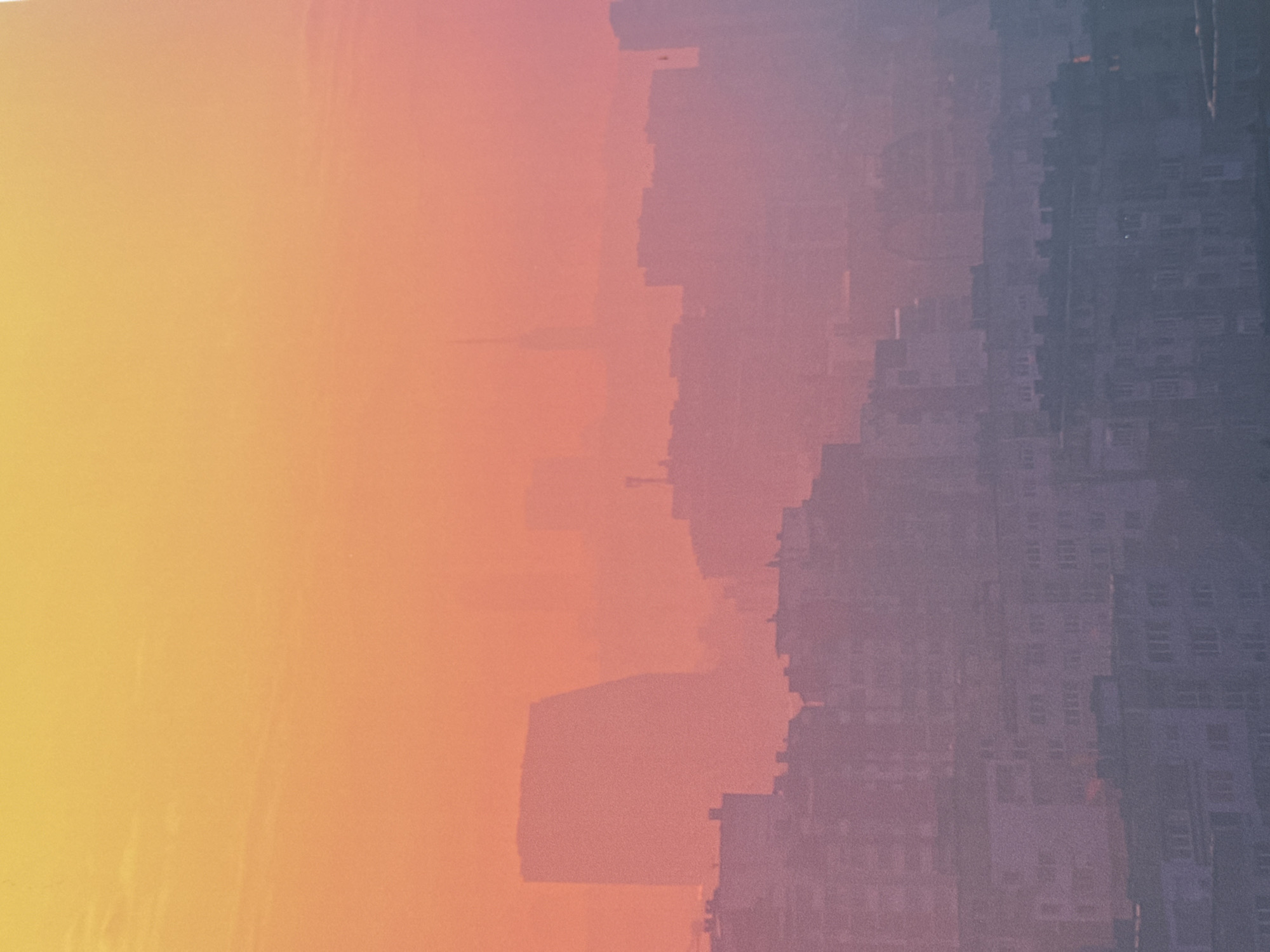 The image depicts an upside-down cityscape under a gradient sky. The sky transitions from yellow to orange to pink. The cityscape consists of densely packed buildings, primarily residential blocks with multiple windows, creating a patterned texture. The buildings fade into a hazy, greyish-purple color. The image is oriented in reverse with the ground at the top and the sky at the bottom. The location is unidentified and does not include known landmarks. There are no discernible people or specific objects within the city buildings. The overall environment is urban, with a hazy atmosphere.