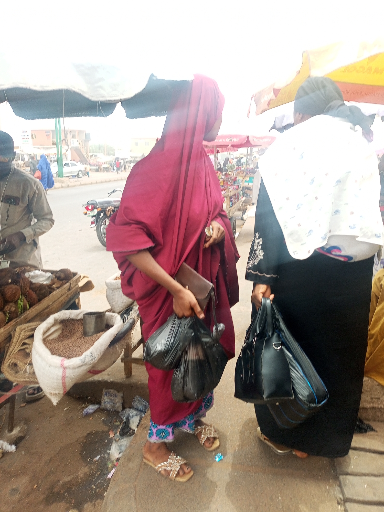 This image depicts a bustling outdoor market scene in Tempure, Nigeria. In the foreground, two women are captured mid-activity. On the left, a woman wearing a deep maroon head covering and flowing traditional attire, with patterned trousers underneath, holds several black plastic bags and a small brown wallet/purse. She stands in embellished sandals next to a market stall. This stall features a wooden crate filled with coconuts, some showing new sprouts, and a large white sack containing what appears to be seeds or grains, possibly groundnuts, with a metal cup resting inside.
To the right, another woman is seen from behind, dressed in a black abaya with embroidered sleeves and a white patterned head covering. She carries a black handbag and wears sandals. A man in a light-colored tunic and cap is partially visible on the far left, seemingly tending to another stall.
The background shows a lively street with a motorcycle parked and other vehicles, as well as various buildings under a bright, possibly overcast or hazy sky. Market stalls are shaded by canopies and umbrellas; one red canopy in the upper right features partially legible, mirrored text that appears to be "MAGGI." The ground is a mix of dirt and pavement, with some scattered debris. The scene conveys the daily commerce and vibrant atmosphere of a typical West African street market during daytime.