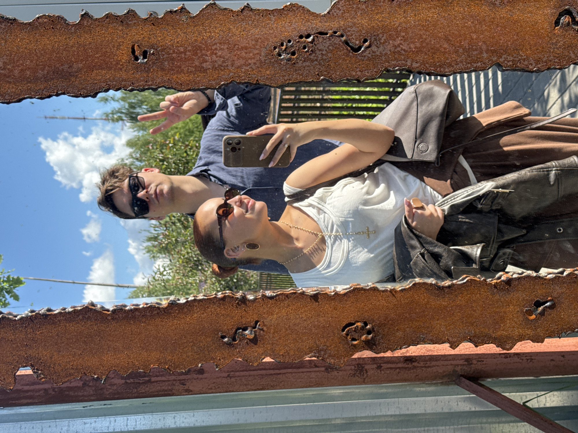 A mirror selfie shows a couple in what appears to be an outdoor setting. A man is on the left, wearing sunglasses and giving a peace sign. A woman is on the right, wearing sunglasses, a white top, and jewelry, and holding a phone. The mirror has a rusted metal frame. The background shows greenery, a building, and a bright sky with clouds. While it is impossible to determine the location without further information, the prompt suggests it is in Izhevsk, Russia.