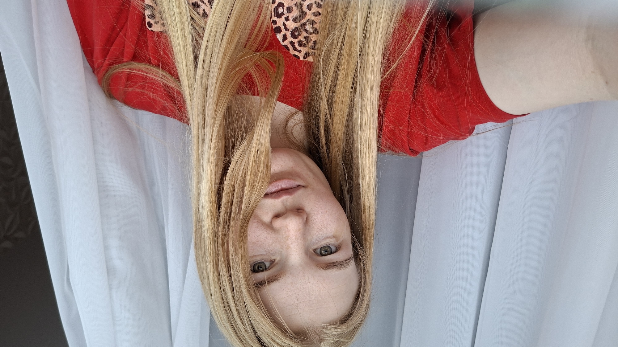 A medium shot selfie shows a young woman with blonde hair and fair skin, wearing a red t-shirt with a leopard print design. She is looking at the camera with a neutral expression. Her hair is long and straight, falling around her shoulders. The background consists of white sheer curtains, suggesting an indoor setting. There are no identifiable landmarks or clues in the image that would indicate a specific location in the city of Chaikovsky, Russia. The lighting is natural, likely from a window.
