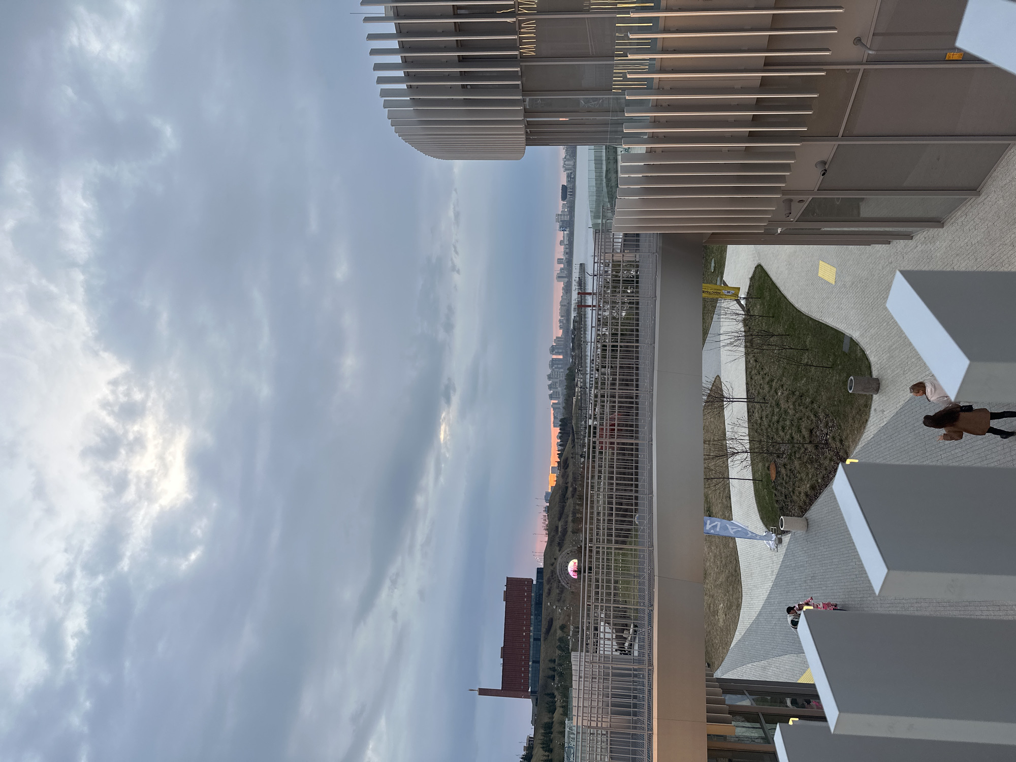 The image shows an elevated view in Kazan, Russia. The sky is overcast with light shining through the clouds. A distant cityscape with a hint of orange is visible on the horizon, including the Kazan Kremlin and Qolşärif Mosque. Below, there is a multi-level building with modern architectural features like metal grating and slatted overhangs. There is a landscaped area with grass and paving, and several people are walking along the pavement, including a child and an adult. There are also several street lights in the view.
