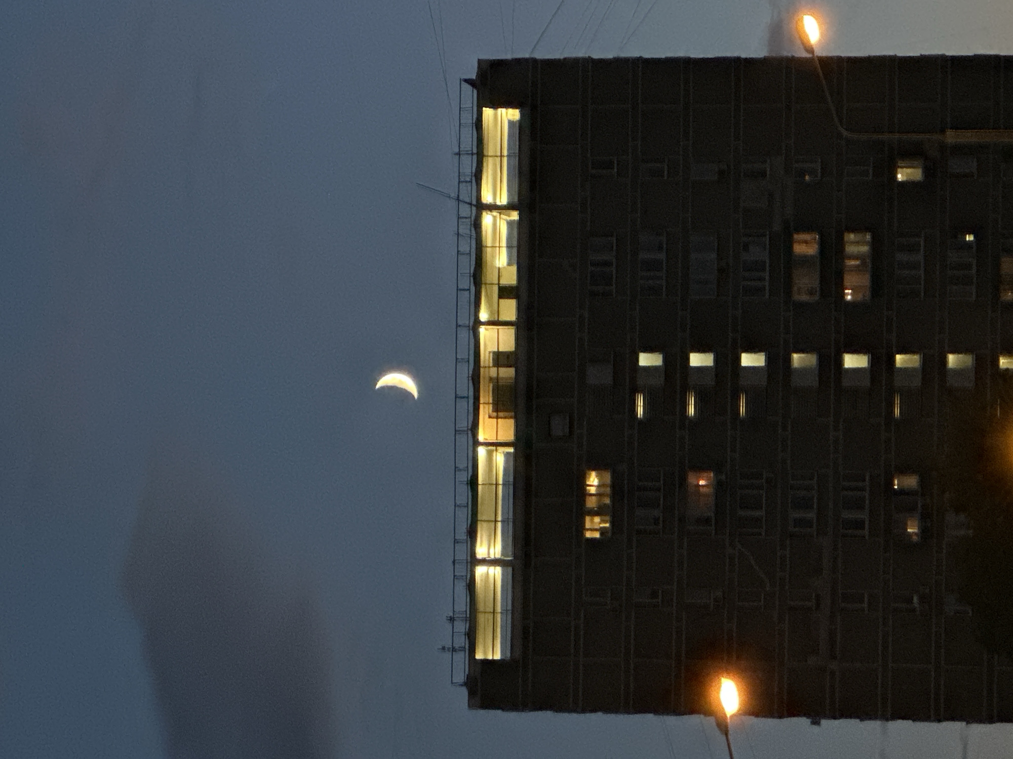The image shows a nighttime view of a large, multi-story apartment building in Moscow, Russia. The sky is a deep twilight blue with a faint crescent moon visible in the upper right quadrant. A dark, wispy cloud is present in the upper left corner. The building itself is dark, with numerous windows, many of which are illuminated with warm yellow light, suggesting occupied apartments. The top of the building has a well-lit, glass-enclosed section. A streetlamp with an orange glow is visible on the right side of the frame, and another is partially visible on the left. Power lines stretch across the sky. The scene depicts a typical urban residential environment at dusk or early night.