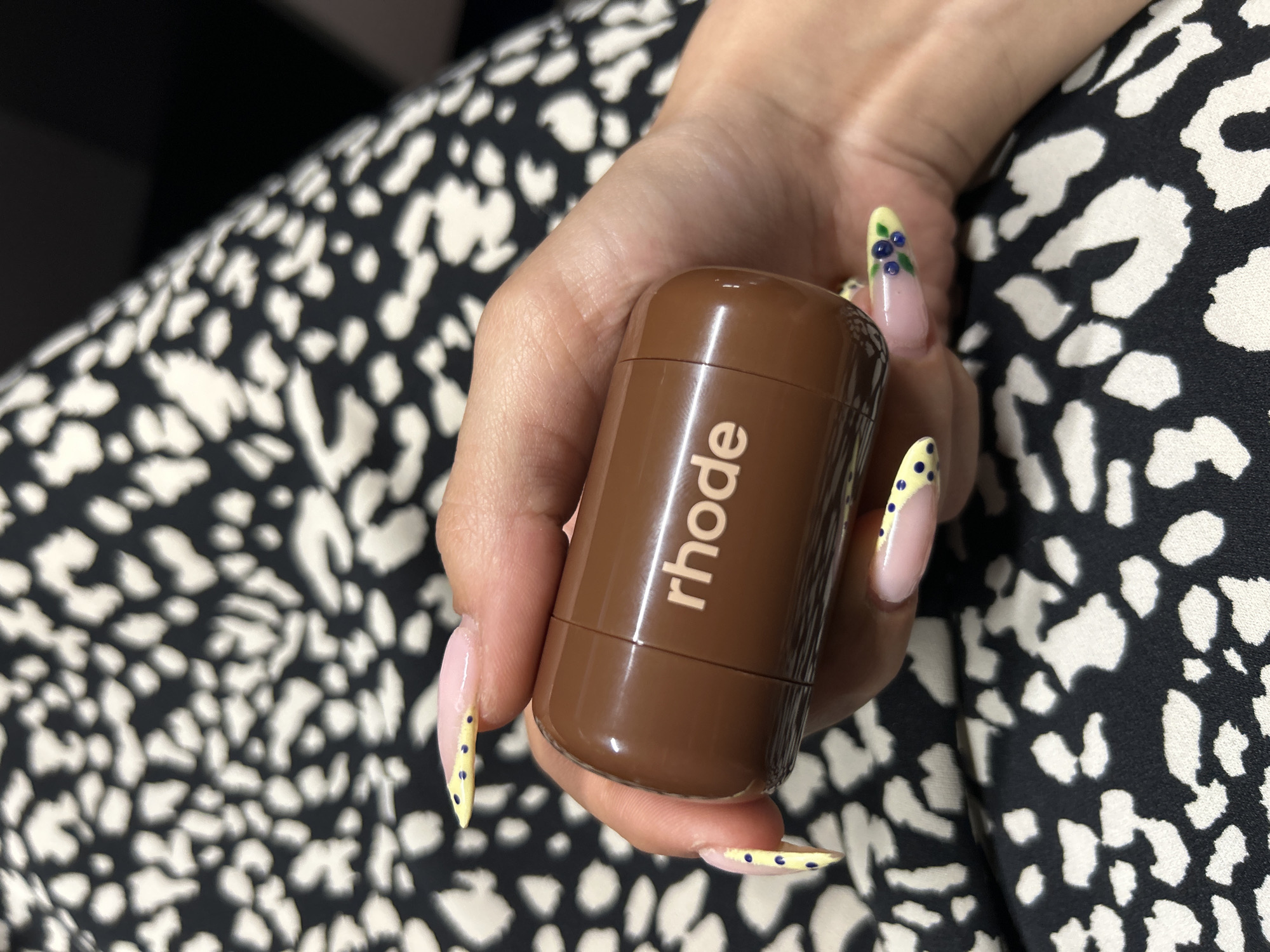 An image shows a person's hand holding a brown cylindrical object with the word "rhode" printed in white. The background consists of a black and white patterned fabric. The person's fingernails are painted with a light pink color and decorated with yellow tips, blue dots, and a small blueberry design on one nail. The scene is indoors, and there is no discernible information to suggest a location in Tbilisi, Georgia.