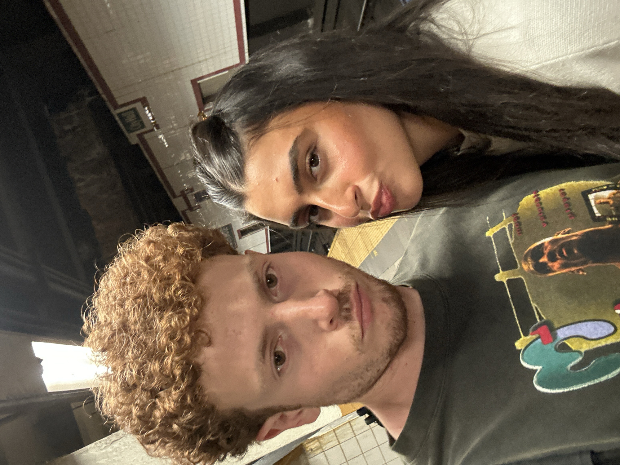 A selfie taken inside a New York City subway station features a male and a female. The male, positioned in the foreground, has light skin, curly reddish-brown hair, and a light beard. He is wearing a dark-colored t-shirt with a faded graphic. The female, with darker skin and long, dark hair, is leaning into the shot from behind. The background shows the typical tile walls and signage found in NYC subway stations.