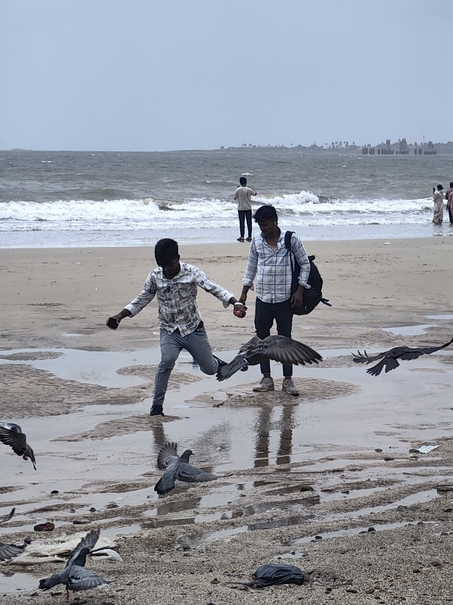 This image depicts a beach scene in Mumbai, India, on a cloudy day with choppy waves. In the foreground, two young men are interacting with pigeons on the wet sand. The man on the left, wearing a patterned shirt and jeans, appears to be in motion, possibly playing or feeding the birds. He is holding a small red object, likely a fruit or ball, and a phone. The man on the right, in a checkered shirt and jeans, is standing with a backpack slung over his shoulder, observing the scene. Several pigeons are in flight or gathered on the sand, attracted by the activity. In the background, other beachgoers are visible, as well as distant structures and a treeline suggesting an urban coastline. The wet sand, shallow pools of water, and overcast sky create a somewhat muted and atmospheric setting.