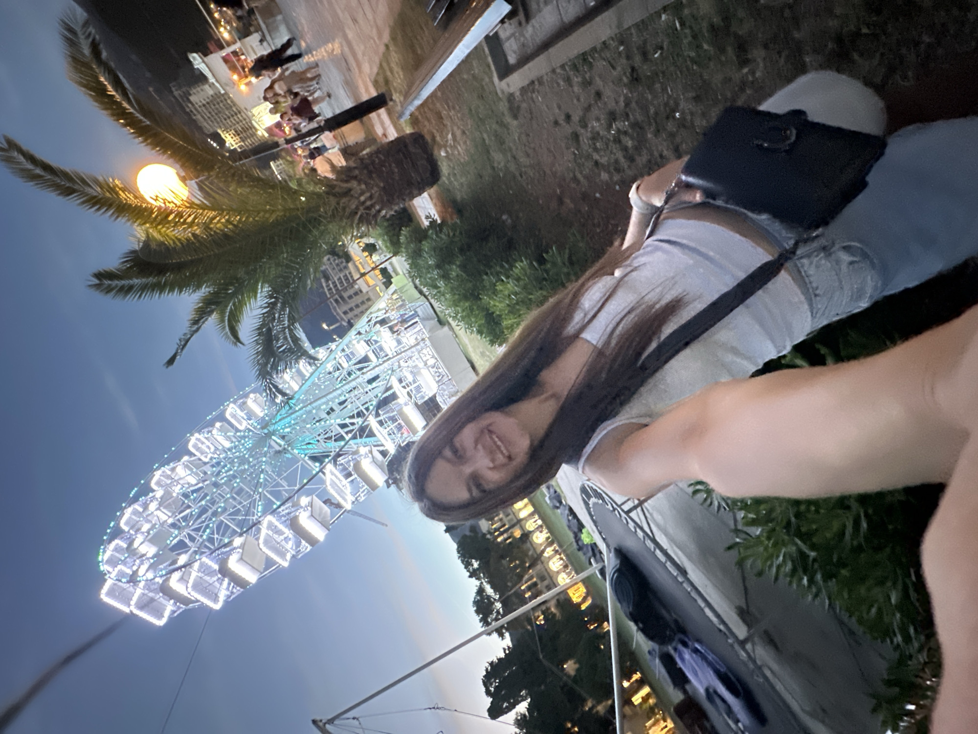 In Budva, Montenegro, at dusk, a woman with long, dark hair takes a selfie in front of a brightly lit Ferris wheel. She is wearing a white tank top and light-colored jeans. A dark purse hangs from her shoulder. Behind her, a palm tree with an illuminated light fixture stands prominently, and further back, a crowd of people walks along a promenade. Buildings and more trees are visible in the background, and the sky is a deep blue, indicating the transition from day to night. A trampoline is visible in the lower-left portion of the image.