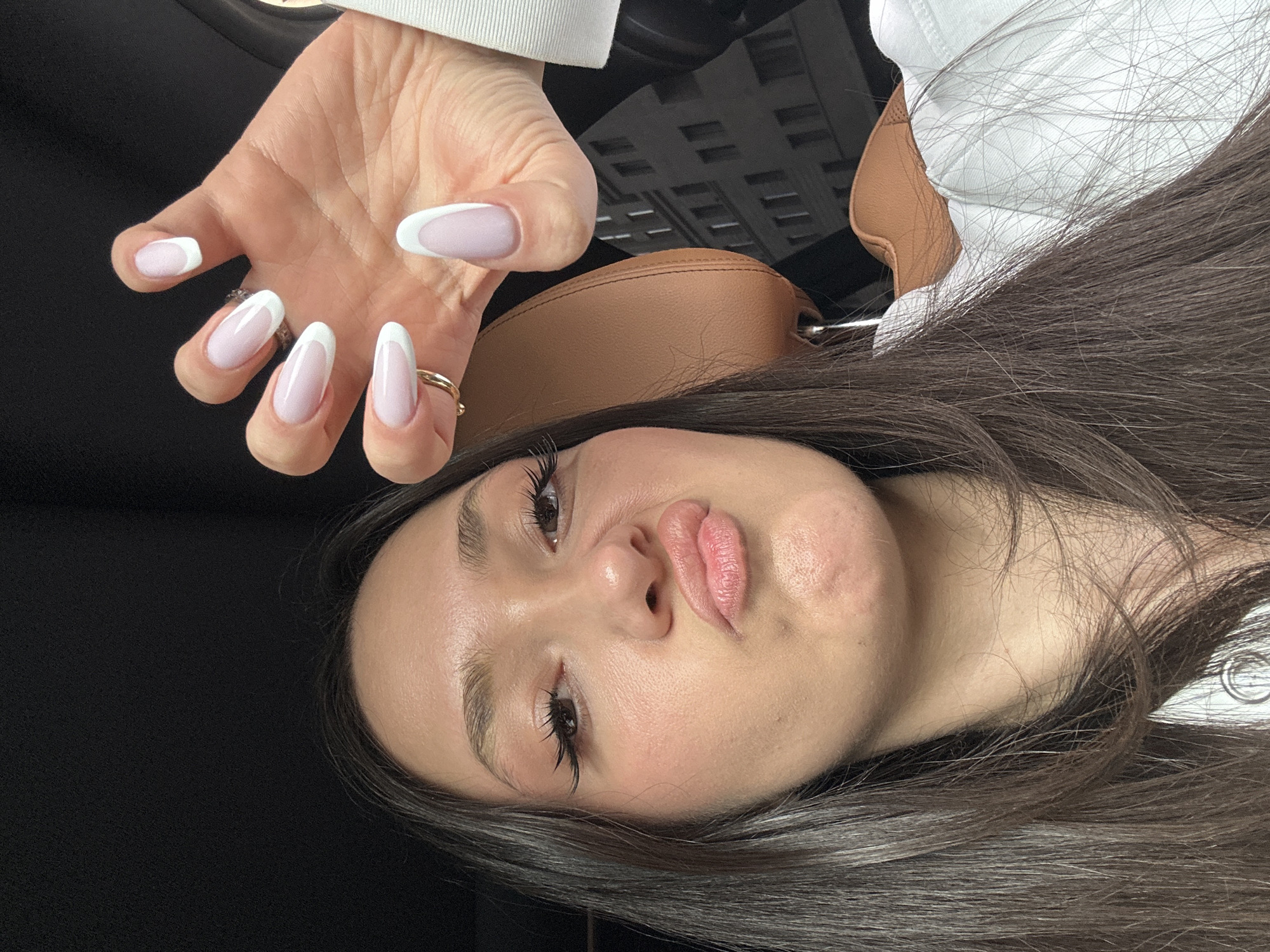 A selfie-style shot captures a fair-skinned woman with brown hair, posed with her face angled towards the camera and her hand raised above her head. The woman has dark eyelashes, full lips, and a clear complexion. She wears a white garment. Her hand displays almond-shaped nails with a white French manicure. On her finger is a gold ring.
In the background, a brown leather headrest of a car seat and parts of a building visible through the car window. The environment appears to be inside a car.
There is no explicit information that indicates the location in Yerevan, Armenia.