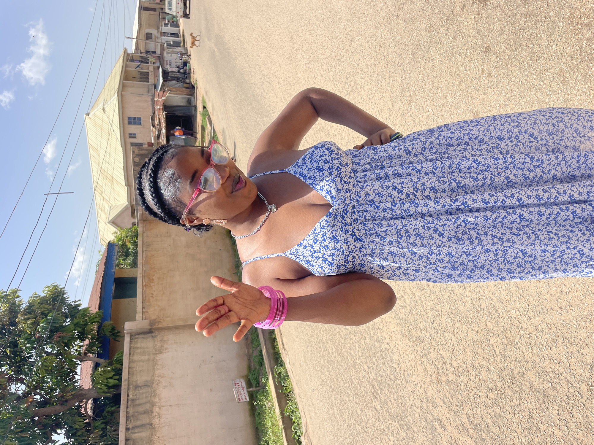 A woman with braided hair and pink-rimmed glasses stands on a paved street, posing for a photo. She wears a blue and white floral print dress and has several pink bracelets on her left arm. Her right arm is bent at the elbow, and her hand is open, palm facing the camera. The scene appears to be outdoors in a street lined with buildings and some vegetation. The sky is visible, with clouds and what appear to be telephone wires overhead. Sunlight suggests it is daytime. In the background, there are structures that look like shops or houses, and a dog is visible on the street. The overall setting suggests a residential or commercial area in Jos, Nigeria. A small, weathered sign with some text is visible near the bottom left of the image, but the text is not legible.
