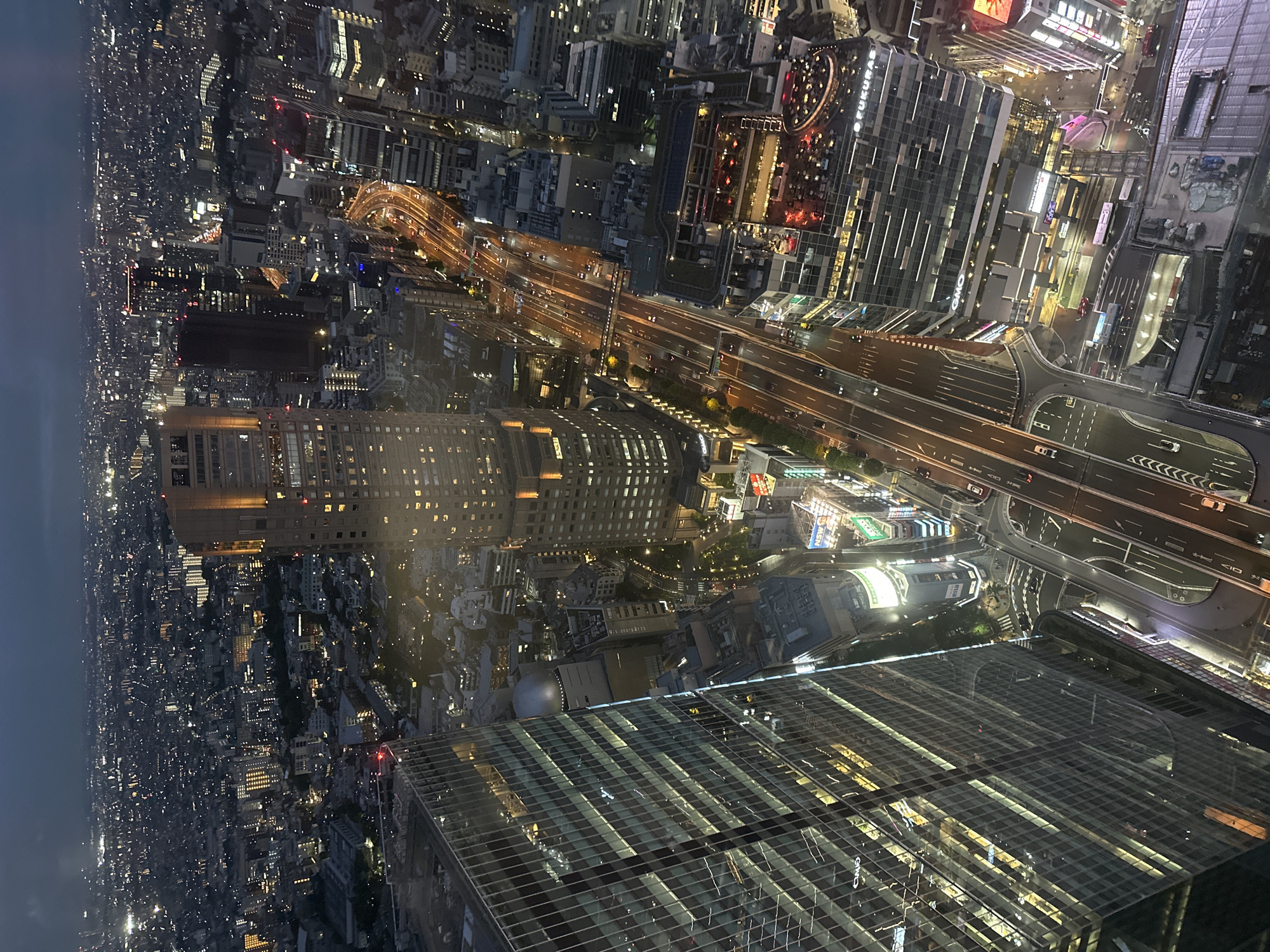 This is an aerial nighttime view of Shibuya, Japan, focusing on illuminated cityscapes. The camera is positioned high, likely within a skyscraper, offering a vantage point that reveals intricate urban details. In the foreground, the reflective glass roof of the building the camera is located in is visible.
The scene is dominated by dense urban development. Numerous high-rise buildings stand densely packed together. Interlaced highways crisscross the area. Numerous orange-colored lights indicate car traffic.
Illumination is plentiful, with buildings lit up by interior lights. The overall impression is a sprawling, vibrant urban environment after dark.