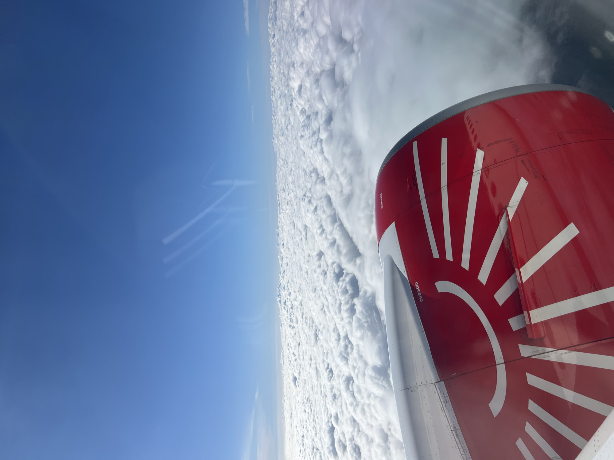 A tilted view from an airplane window shows a red and white tailfin with a sunburst design. Above the clouds, clear blue sky extends to the left. The location is presumed to be in the sky, above an extensive layer of clouds.