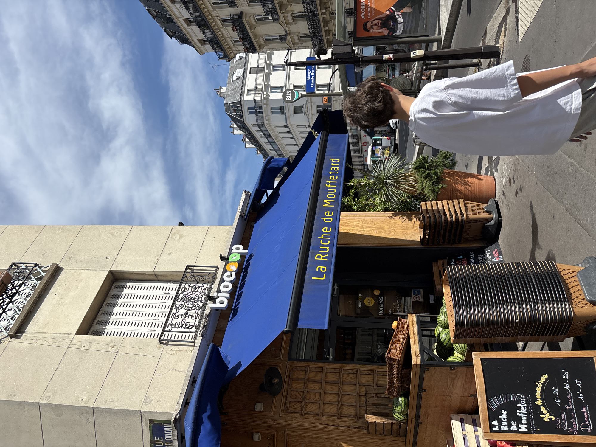 A street-level, medium shot captures a view of a Biocoop store in Paris, France, on a sunny day. The store, named "La Ruche de Mouffetard," has a bright blue awning and wooden storefront. A young person in a white t-shirt is walking away from the camera, towards the right side of the frame. In front of the store, there are displays of watermelons, baskets, and stacked shopping carts. A sign with the store's name and hours is visible on the left. The background shows typical Parisian architecture with apartment buildings, a street sign indicating "Monge Claude Bernard," and a bus stop. The sky is partly cloudy with blue patches.
