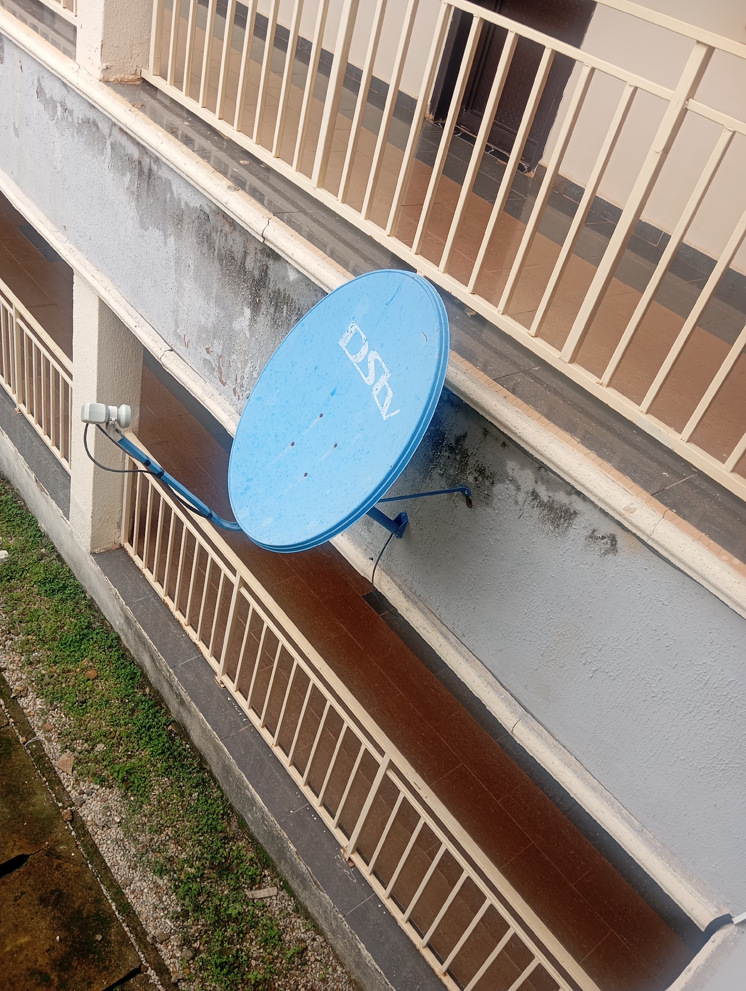 The image shows a satellite dish mounted on the exterior wall of a building, likely in Jos, Nigeria, based on the presence of DStv branding on the dish. The dish is blue and has the DStv logo visible. It is attached to a metal arm extending from a wall that appears to be part of a balcony or walkway. A railing with vertical bars runs along the edge of the balcony. Below the balcony, there is an area with grass and gravel. The building wall has some signs of wear, with patches of discolored or peeling paint. The ground below appears to be tiled. No people are present in the image. The scene depicts a residential or commercial building with common outdoor amenities.