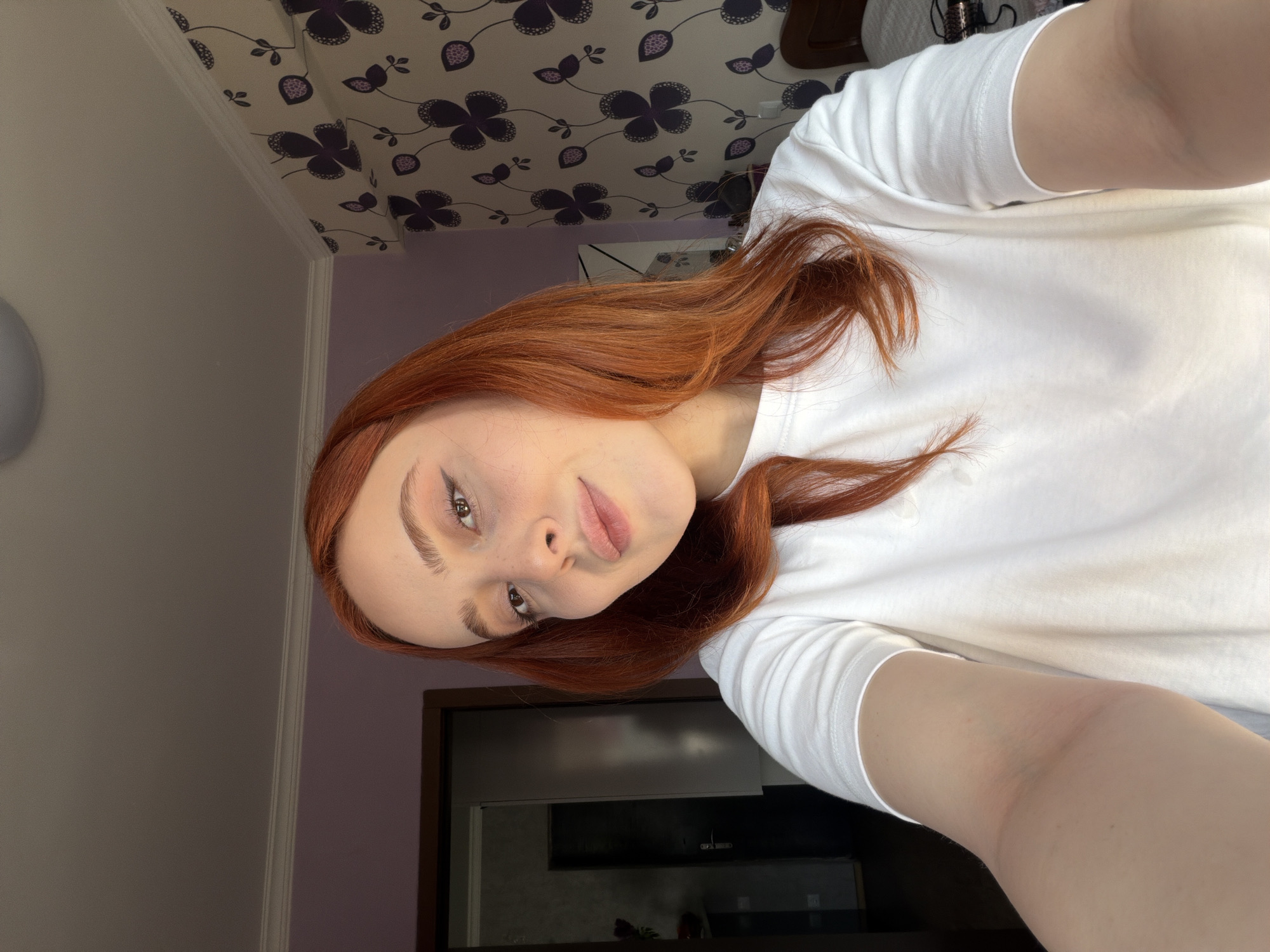 A young woman with auburn hair is taking a selfie. She is wearing a white t-shirt. The background includes a lavender wall, a white wall, a ceiling with a dark purple floral pattern, and a dark piece of furniture. There are no clear landmarks to ascertain a specific location in Tbilisi, Georgia.