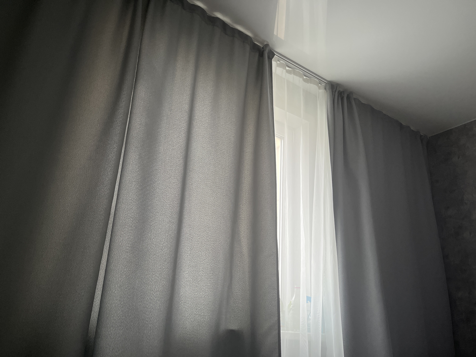 A pair of gray curtains are drawn across a window, with a sheer white curtain visible behind them. The gray curtains are thick and opaque, obscuring most of the view outside. The white curtain is thin and translucent, allowing some light to filter through. The curtains are hung from a metal rod attached to the ceiling. The room appears to be dimly lit, with natural light coming from the window. The wall to the right of the curtains is textured and dark gray. The overall impression is one of a domestic interior, likely a bedroom or living room. The scene is static and there are no people or animals visible. There is no identifiable information within the image to suggest the specific location is Kamskiye Polyani, Russia.