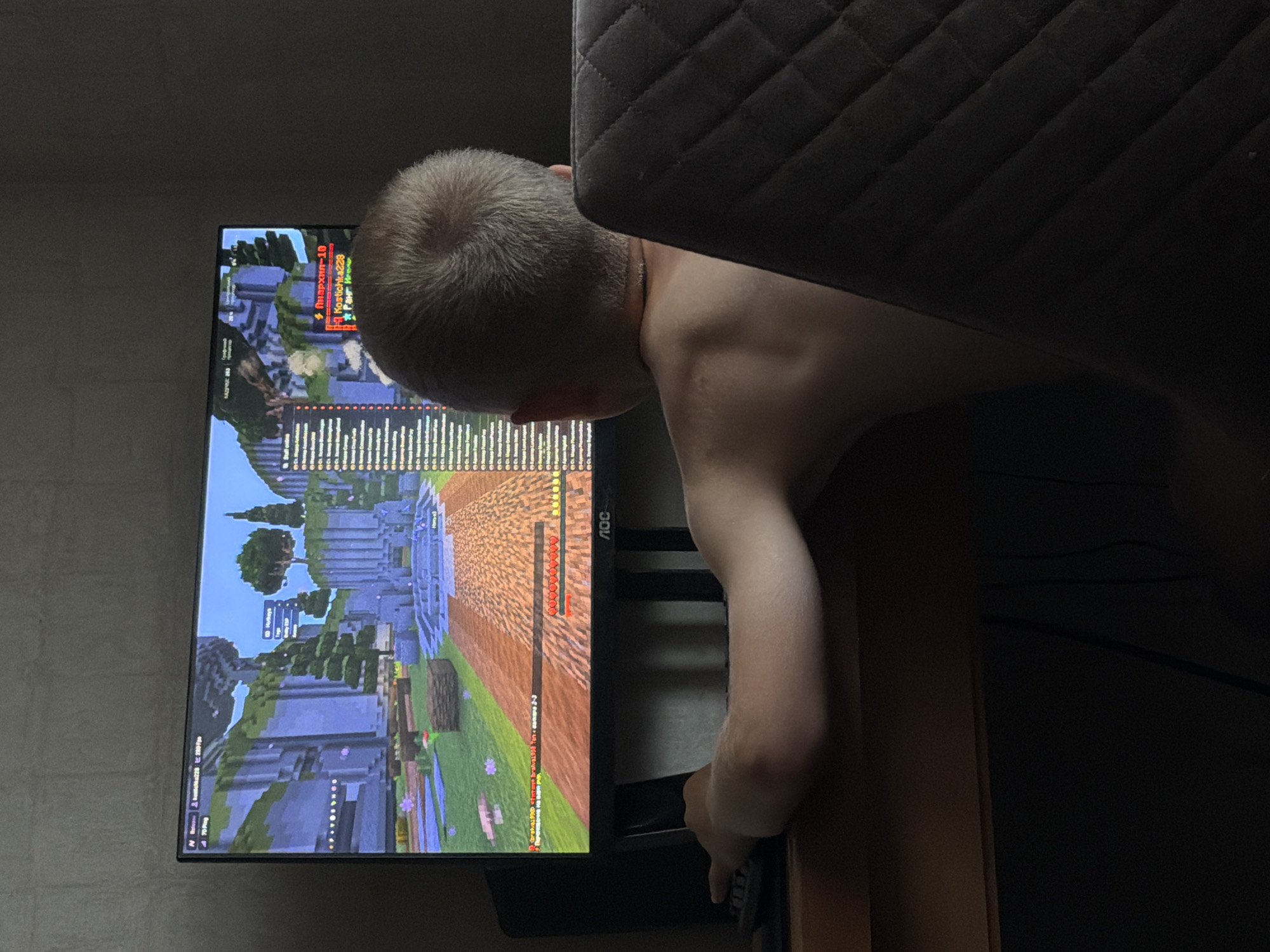 A young boy, seen from behind, sits at a desk playing a video game on a computer monitor. The boy has light brown hair and is wearing a necklace. His left arm is extended, resting on a keyboard. The monitor displays a blocky, pixelated game environment. A chair with a quilted back is visible to the right of the boy. The scene is dimly lit, suggesting it is indoors, possibly at night. There is no visible indication of the city of Vilshany, Ukraine.