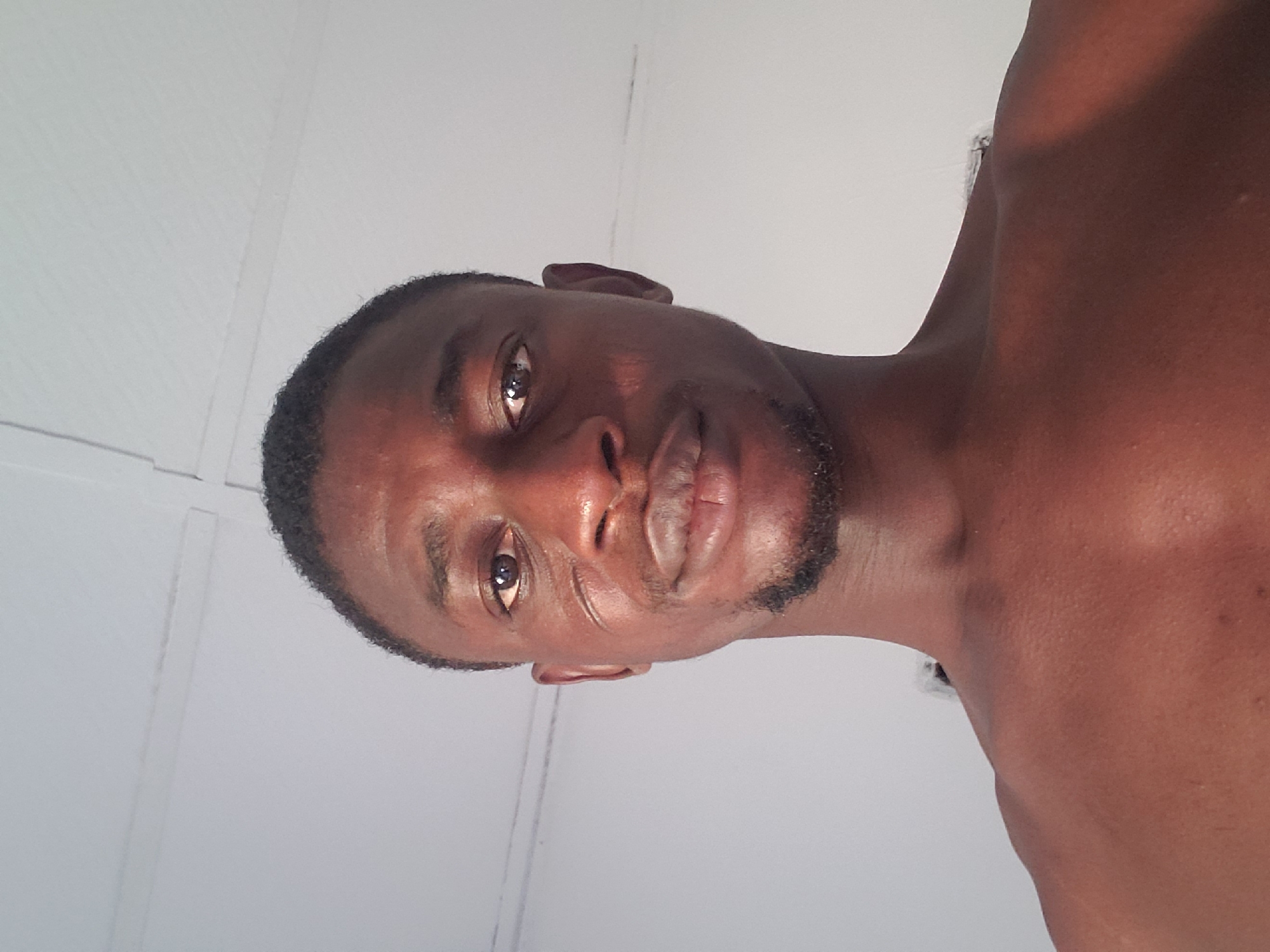 The image shows a medium-close-up of a dark-skinned male, possibly in his late teens to early thirties. He has short, dark hair and a trimmed beard. The background is a plain, light-colored wall. The location, Adonkolo, Nigeria, cannot be verified from the image.