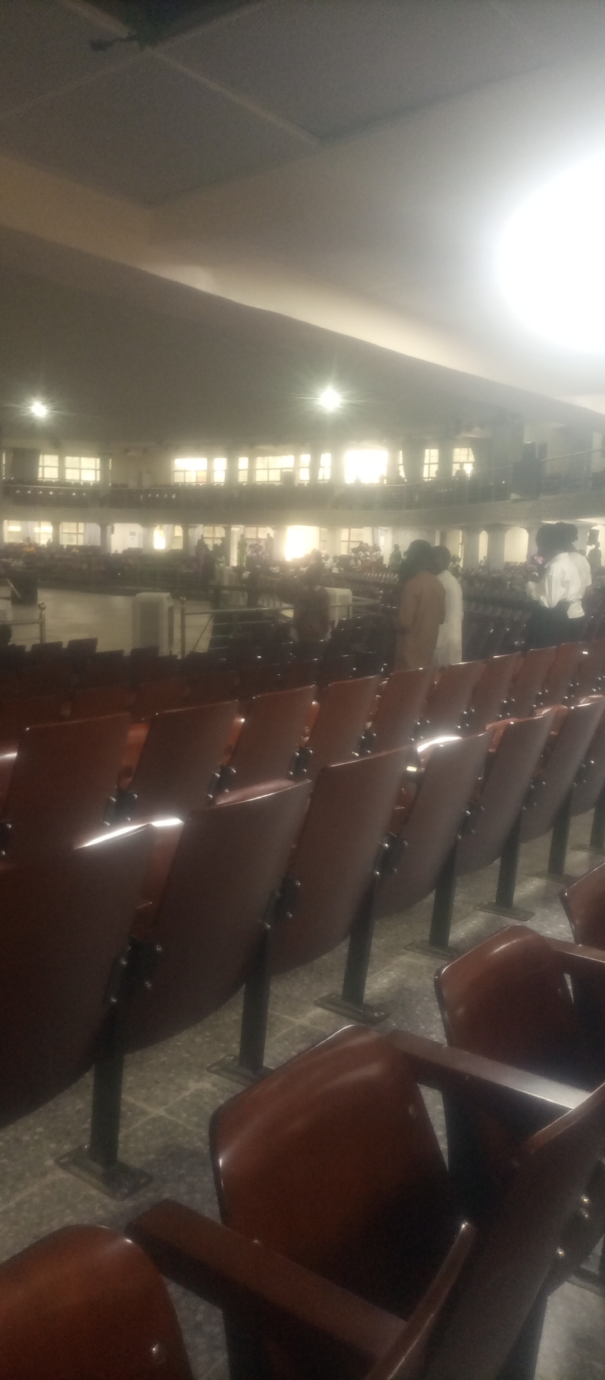 This image depicts the interior of a large hall, likely an auditorium or conference center, in Kuson, Nigeria. Rows of dark brown theater-style seats with metal legs are visible in the foreground, facing a stage or presentation area in the background. The hall features multiple levels with balconies or mezzanines, distinguished by windows and railings. In the background, a gathering of people can be seen, some standing and conversing, while others appear to be seated in the distance. The lighting suggests it is either daytime with bright sunlight filtering through the windows or artificial lighting is in use. The overall atmosphere is that of a public gathering or event.