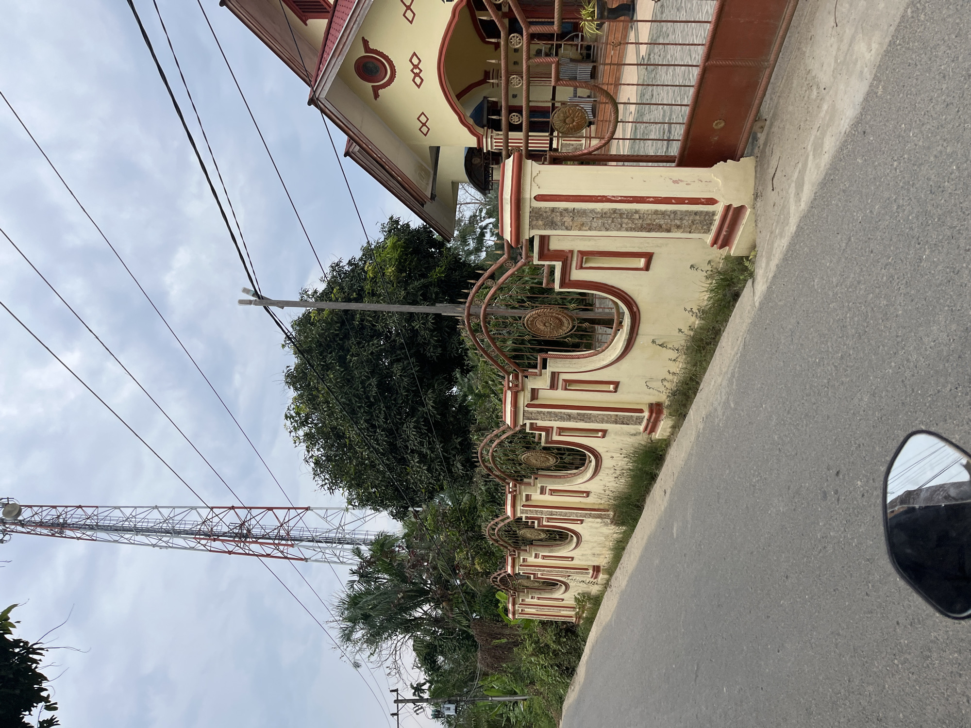 The image shows a residential street scene in Tanahterban, Indonesia, featuring a long, ornate fence along the side of the road. The fence is constructed with alternating sections of light yellow plaster and red-trimmed ornamental pillars, punctuated by decorative metalwork panels. Beyond the fence, a light yellow house with a red roof and red trim is visible. Behind the house and fence, lush green trees and foliage create a dense backdrop. In the distance, a tall, red and white lattice tower, likely a telecommunications tower, stands against a cloudy sky. Overhead, several power lines crisscross the frame. The foreground shows a portion of an asphalt road and the reflection in a motorcycle's side mirror. The scene is devoid of people.