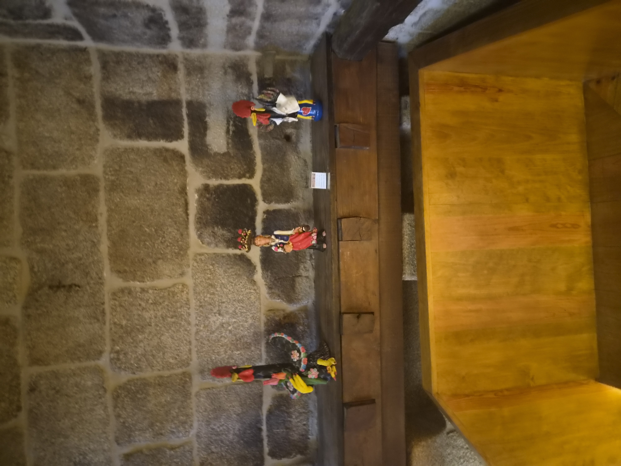 The image shows a rustic interior with a stone wall background and a wooden shelf. On the shelf, there are three decorative figurines. To the left is a black rooster figurine adorned with colorful flowers and yellow accents. In the center, a figurine of a woman in traditional attire stands with a basket on her head. To the right, another rooster figurine, predominantly blue and yellow, is decorated with a red heart. In the foreground, a large, polished wooden structure, possibly a bathtub or a large planter, dominates the lower portion of the image. The overall setting suggests a traditional or craft-oriented environment, possibly a museum or shop, in Barcelos, Portugal, a city renowned for its pottery and folk art, particularly its iconic rooster figures.