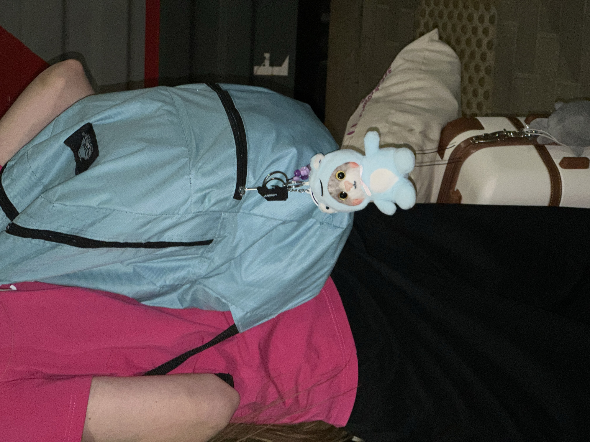 The image shows a person lying down, possibly on a bed. A light blue backpack with black zippers is placed on top of them, partially covering a pink shirt or blanket. A plush toy of a cat in a blue bear costume is attached to the backpack's zipper. To the right, there are bags or luggage with a gray stuffed animal. The environment appears to be dimly lit. No evidence is present to confirm a location in Nizhny Novgorod, Russia.