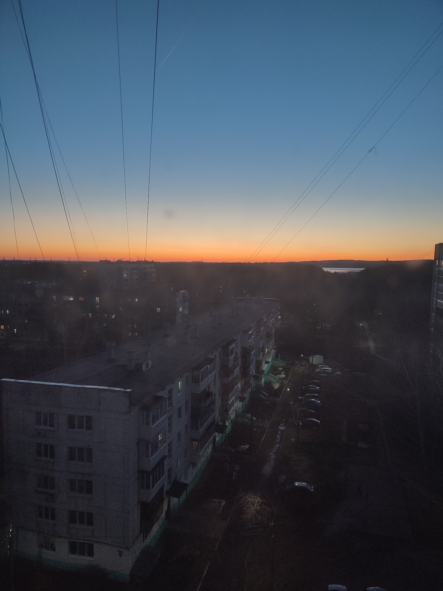 The image shows an aerial view of a residential area in Chaikovsky, Russia, at dusk. In the foreground, there are multi-story apartment buildings with visible windows. The buildings are made of concrete and appear to be aging. A street runs beside the buildings, lined with parked cars. In the distance, the horizon features an orange sky gradient transitioning to a darker blue above. The horizon appears to have a mix of trees and possibly some industrial infrastructure. Overhead, there are visible power lines running across the sky. The overall scene is dark and still.