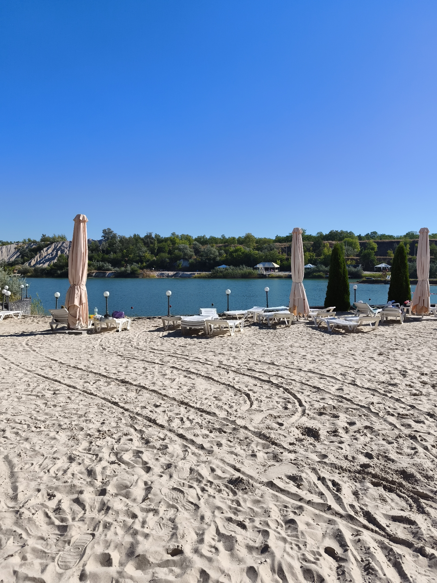 The image shows a sandy beach with a clear blue sky. In the foreground, there are tire tracks and footprints in the sand. In the middle ground, there are several lounge chairs and umbrellas. Behind the beach, there is a body of water and a line of trees. The location is the beach in Katerynivka, Ukraine.